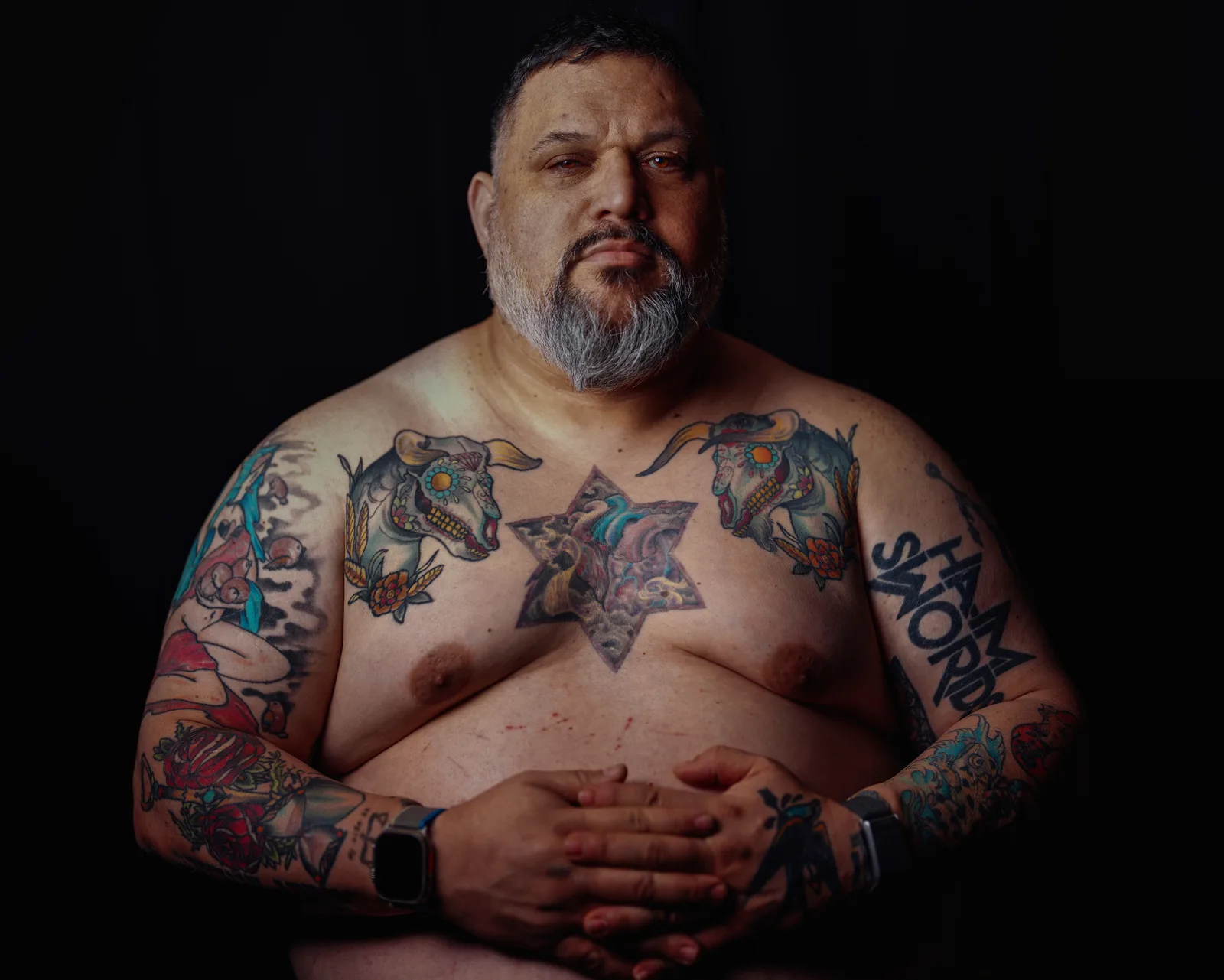 Shirtless torso of a bearded man with colorful chest and arm tattoos, hands folded at his stomach, against black backdrop.