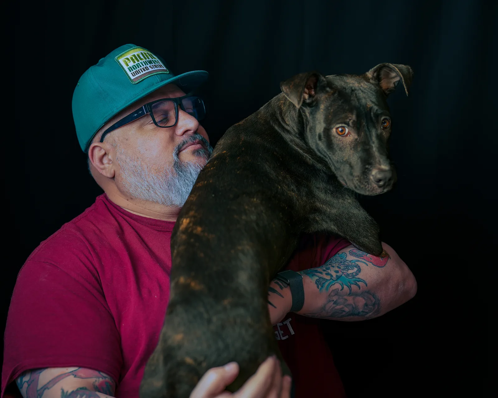 Bearded man in teal Pacific Northwest cap and glasses, eyes closed, resting cheek against a brindle pit mix dog on his shoulder.