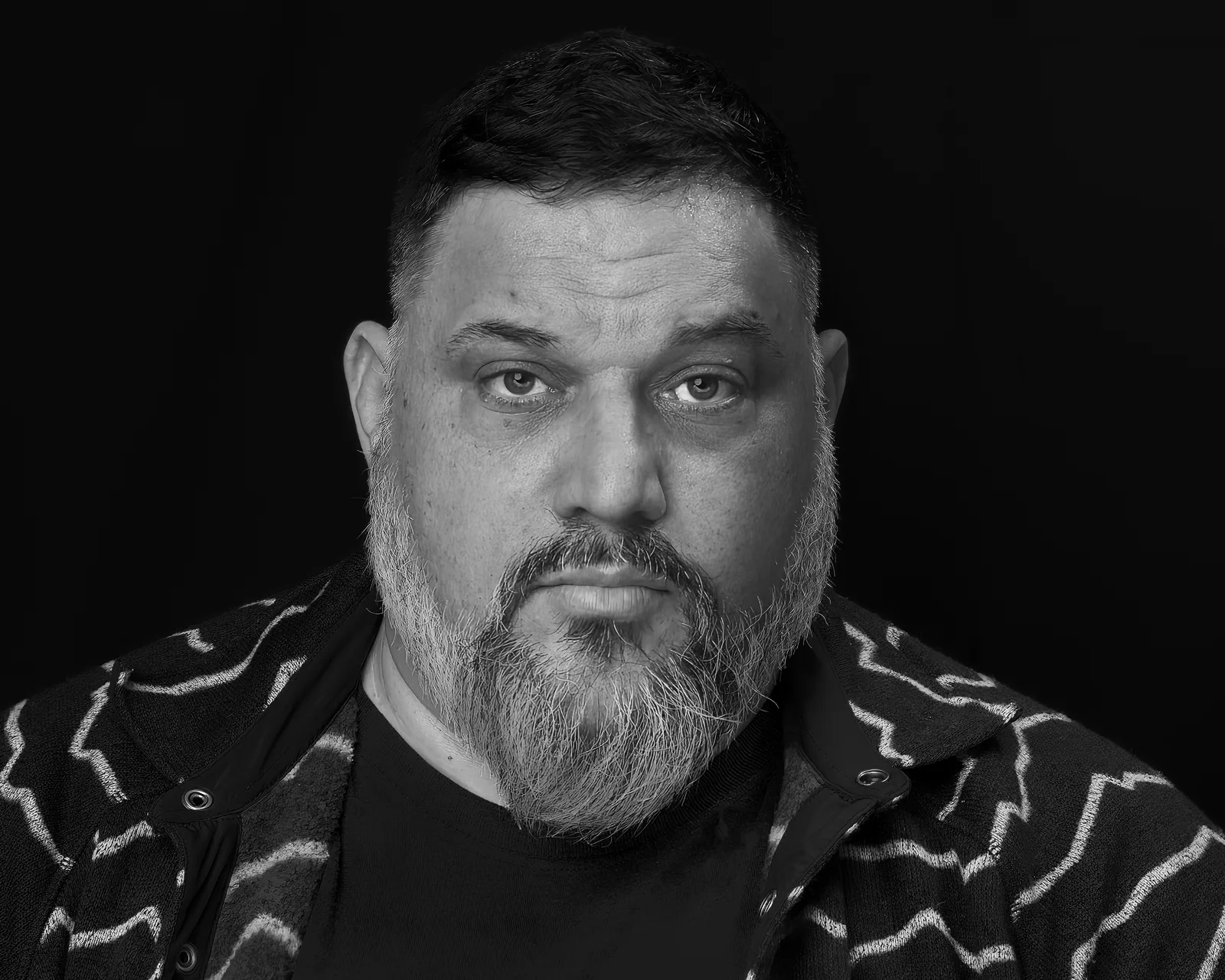 Black-and-white close-up of a bearded middle-aged man with tired eyes, faint sweat on his brow, wearing a patterned jacket against a black background.