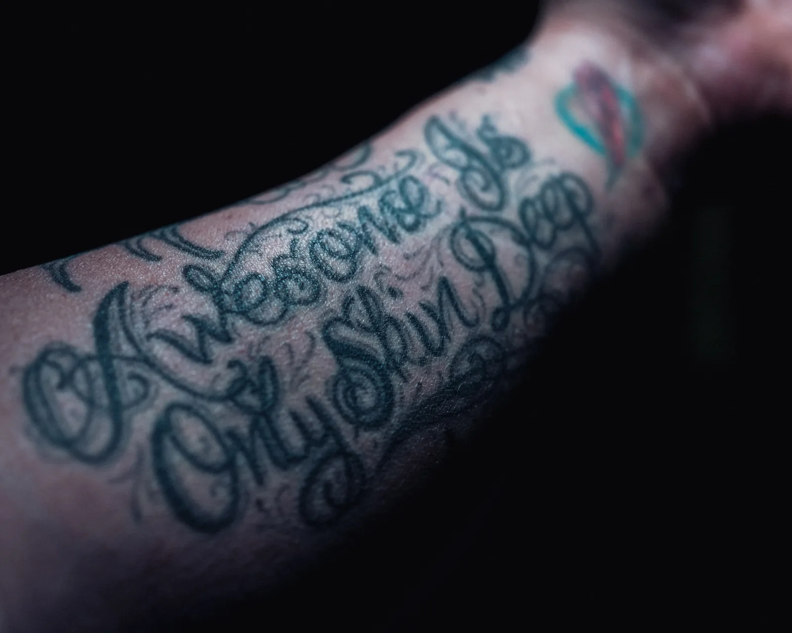 Close-up of a forearm covered in elaborate dark script tattoos, with the words "Awesome" and "Skin Deep" visible against a black background.