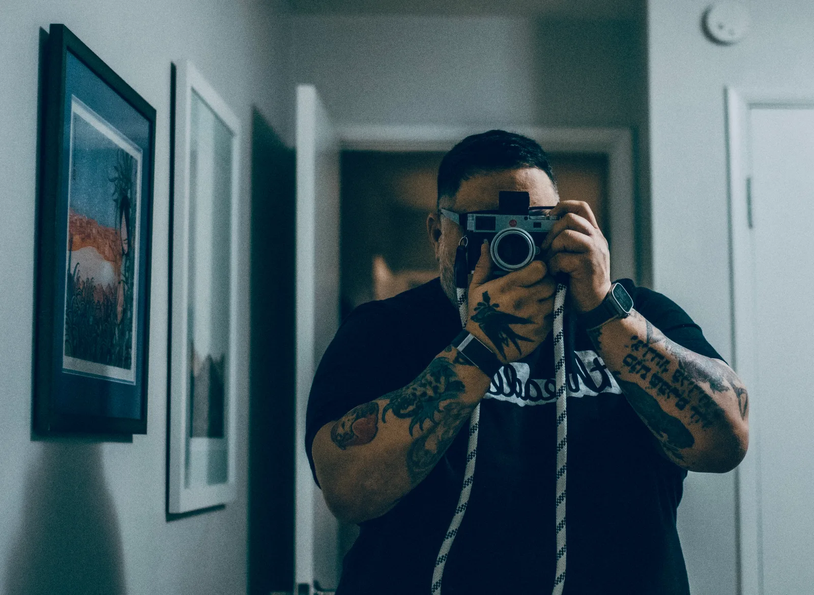 Man with short dark hair and heavily tattooed arms holds a Leica rangefinder camera to his face in a hallway mirror.