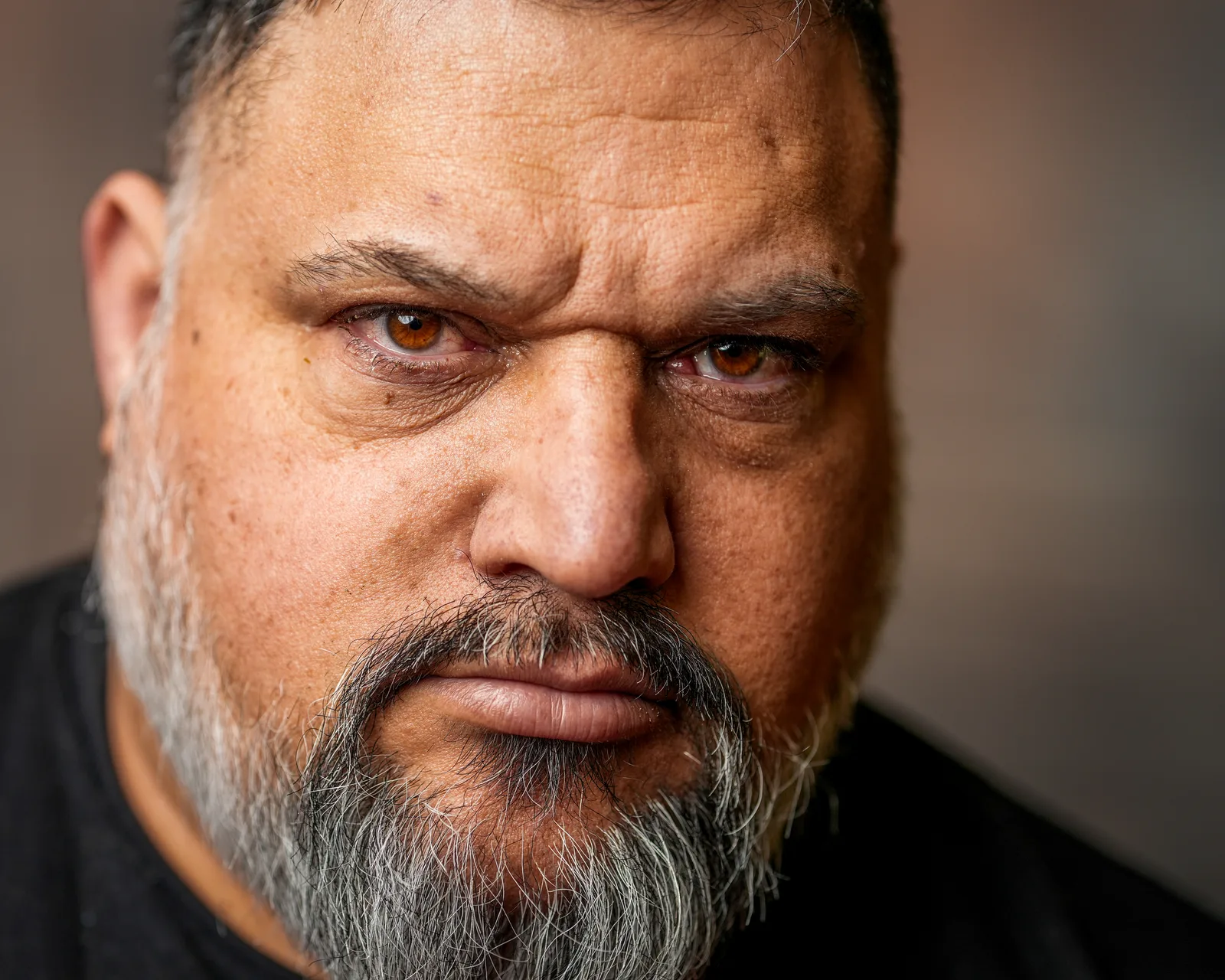 Extreme close-up of a bearded man's face, brown eyes staring directly at camera, faint scar above right brow.