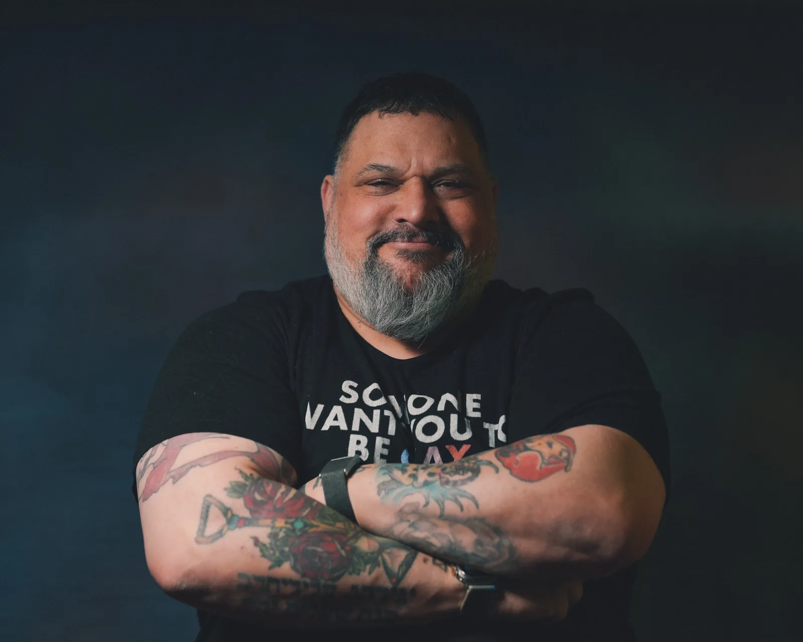 Bearded man with gray-streaked beard, arms crossed showing colorful tattoos, wearing a black graphic t-shirt, slight smile.