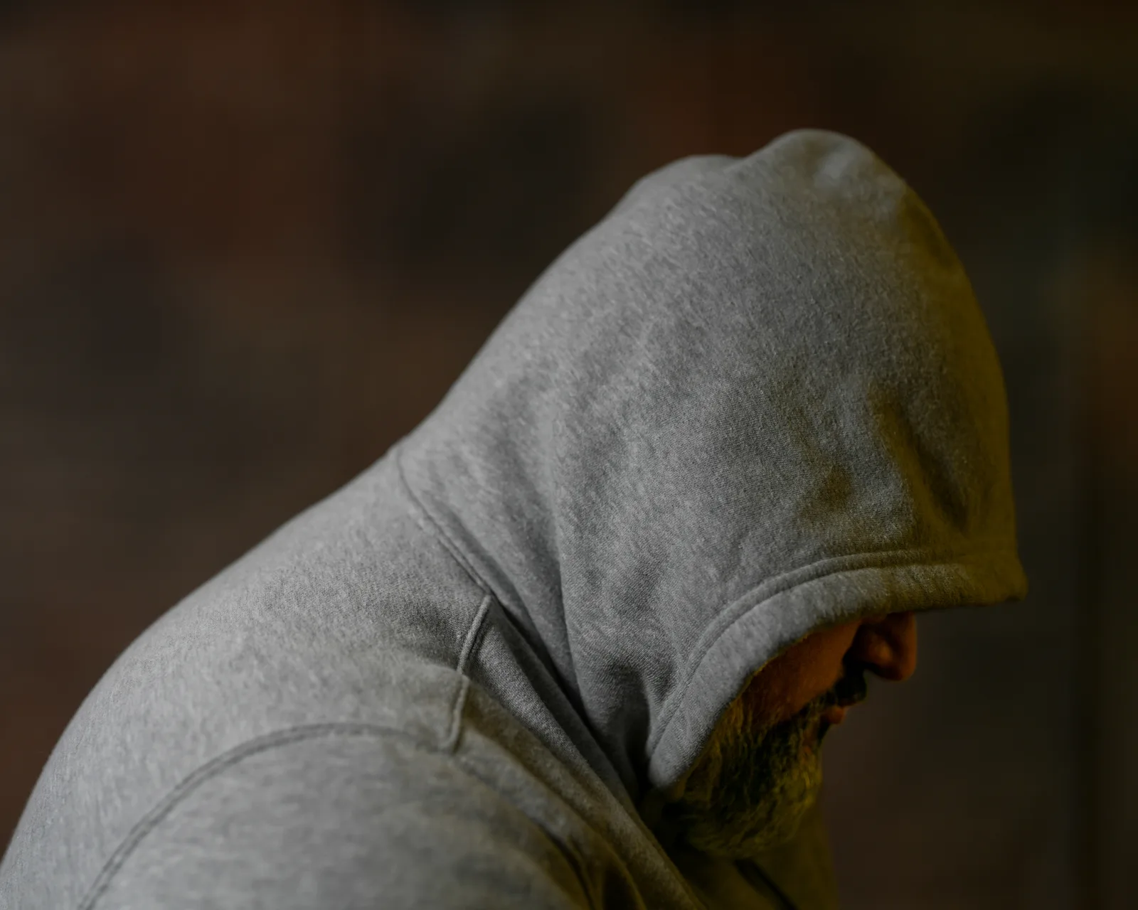 Side profile of a bearded man in a gray hoodie, head bowed, face mostly hidden beneath the hood against a dark background.