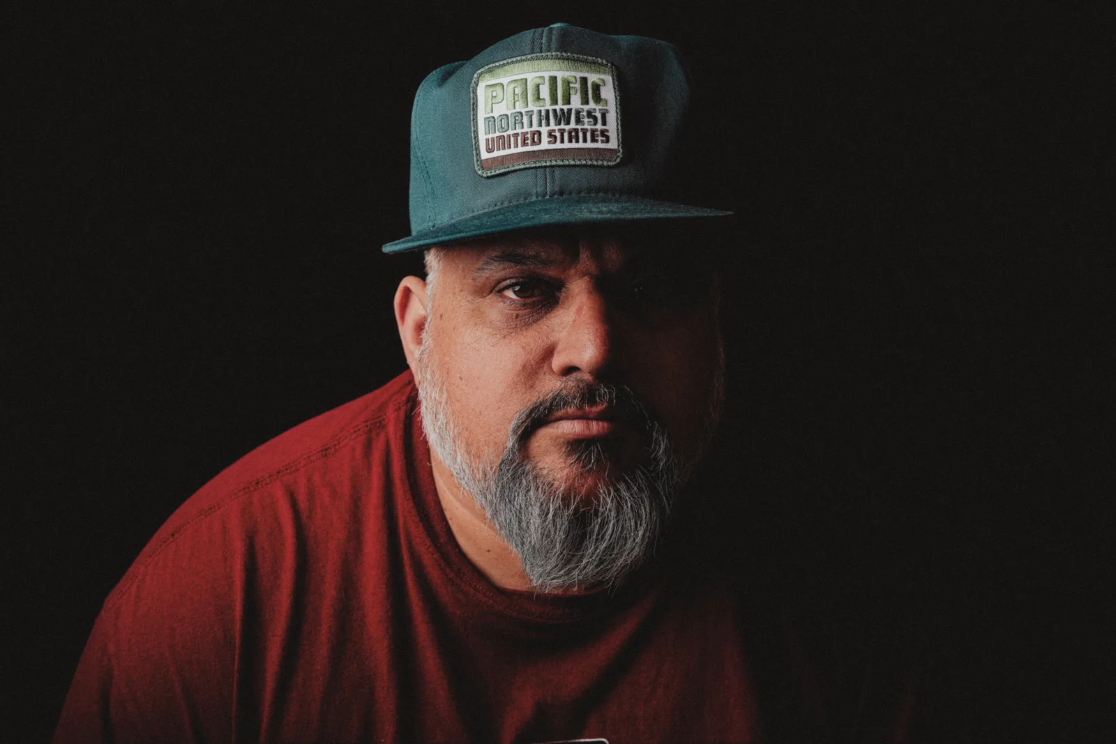 Bearded man in red t-shirt and dark teal Pacific Northwest cap, dimly lit against black background, gazing directly at camera.