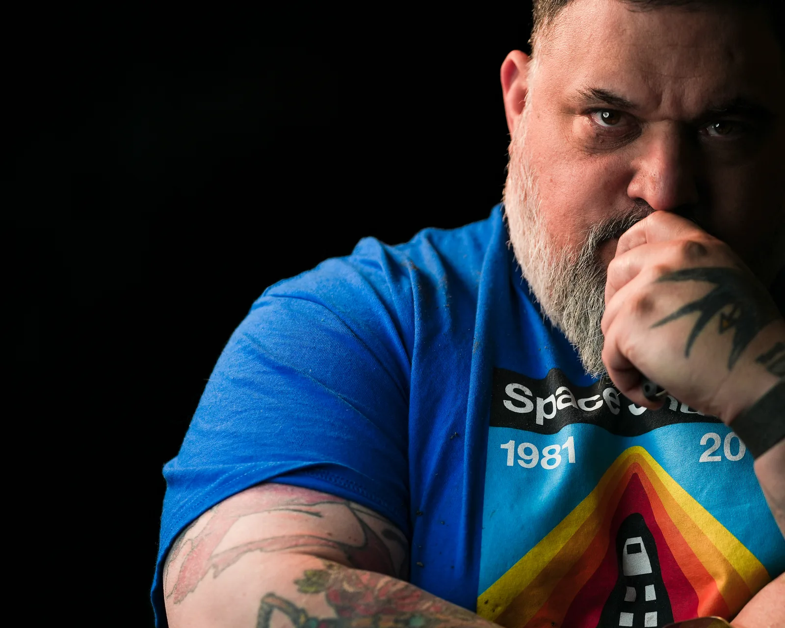 Bearded man in blue t-shirt rests tattooed fist against his mouth, eyes intense, against a black background.