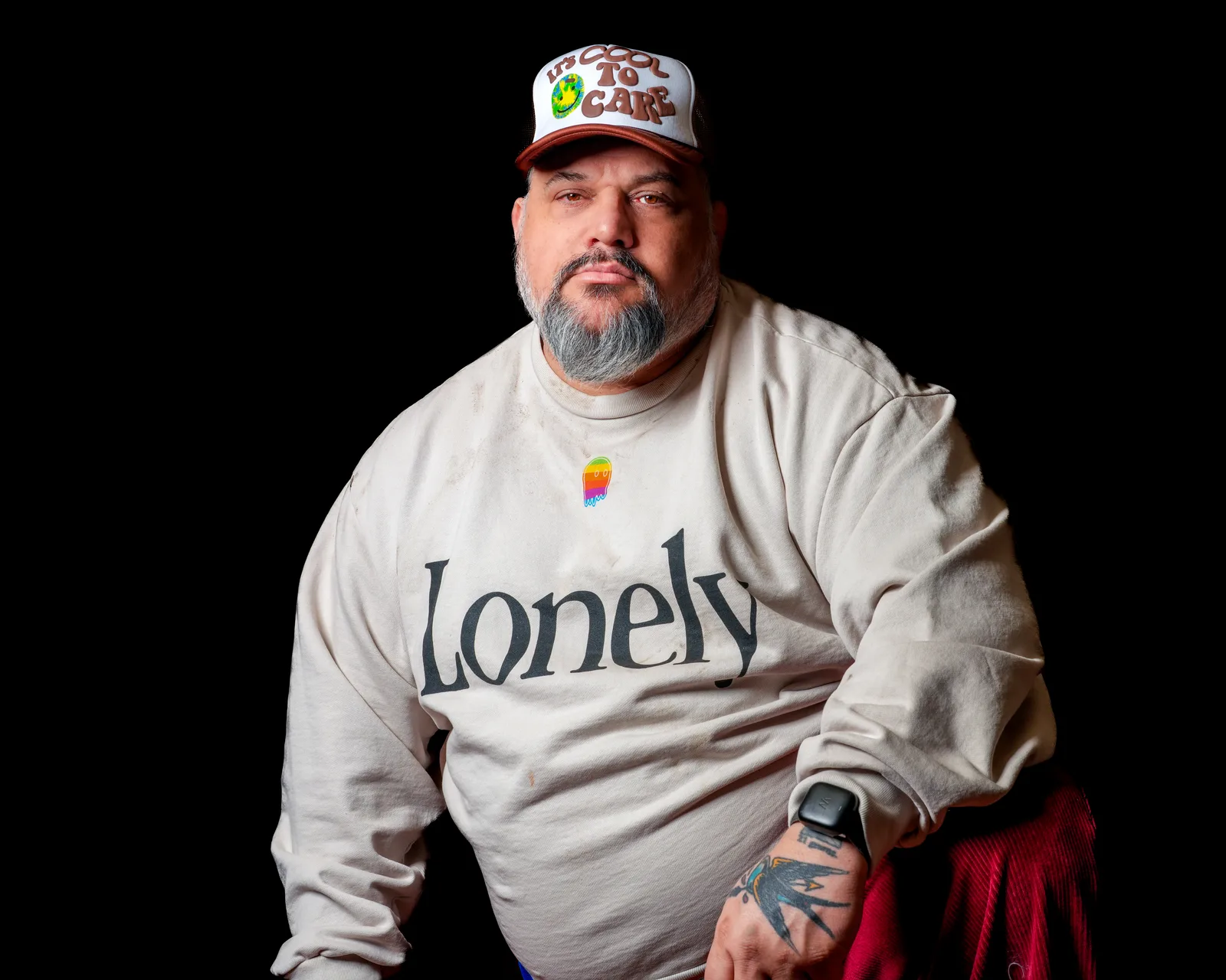 Bearded man's torso in a cream long-sleeve shirt printed with the word 'Lonely', tattooed hand and Apple Watch visible, black background.