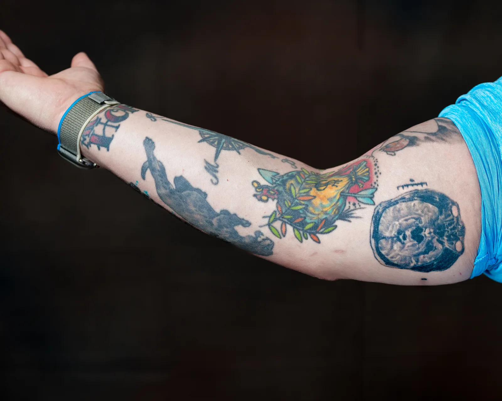Outstretched tattooed arm against black background, showing colorful sleeve with a circular MRI brain scan tattoo near the bicep.