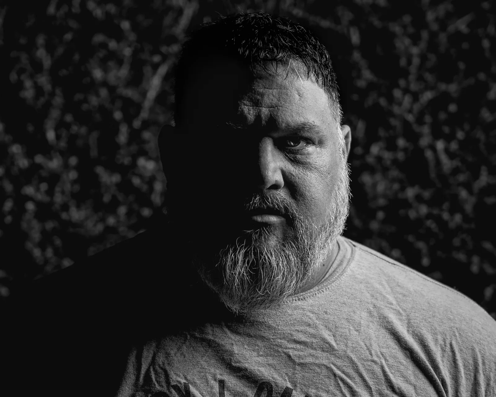 Black-and-white close portrait of a bearded man in a gray t-shirt, half his face in deep shadow, one eye visible.