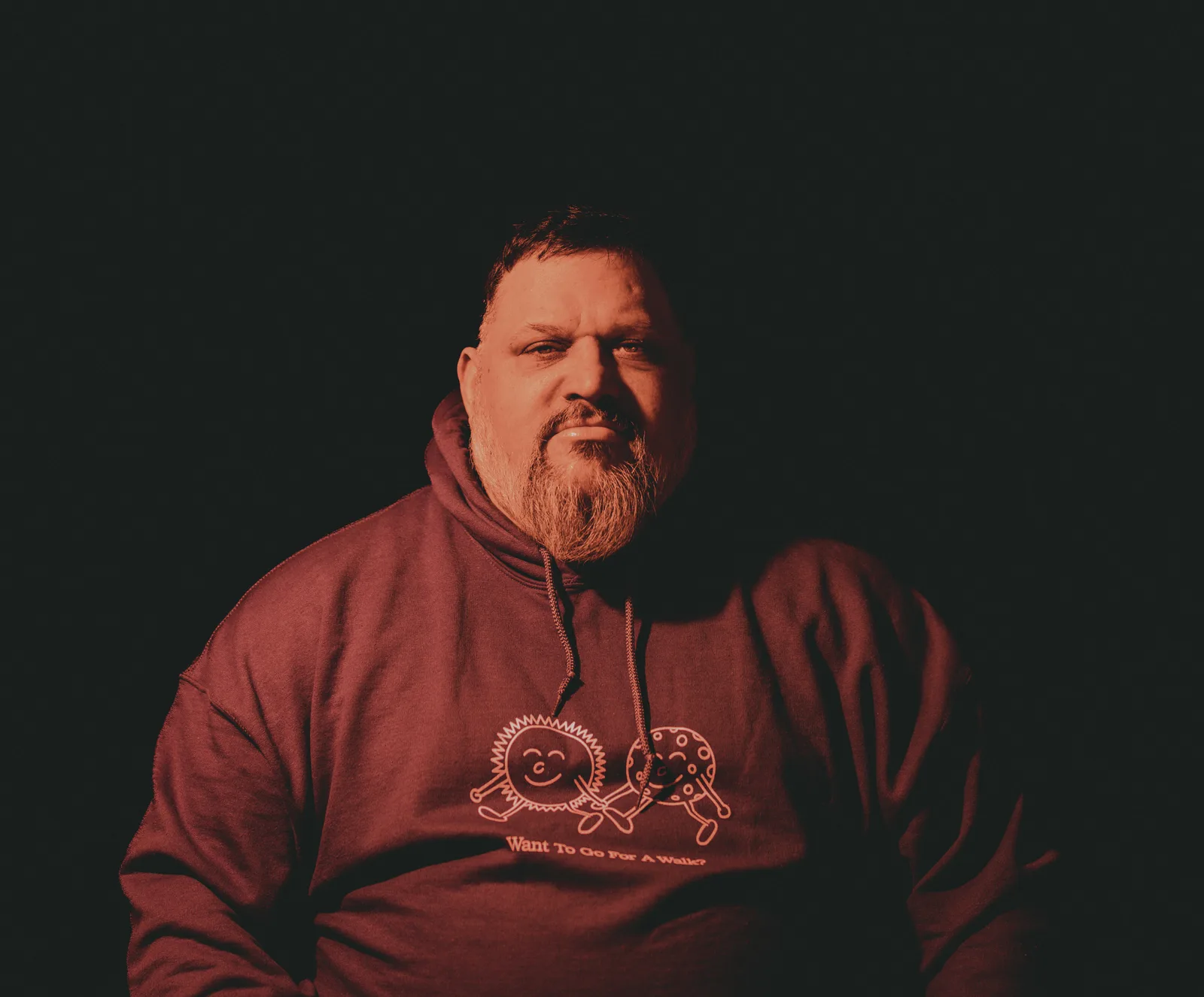 Bearded man with shaved head in maroon hoodie printed with cartoon characters and the words "Want To Go For A Walk?", lit warmly against black.