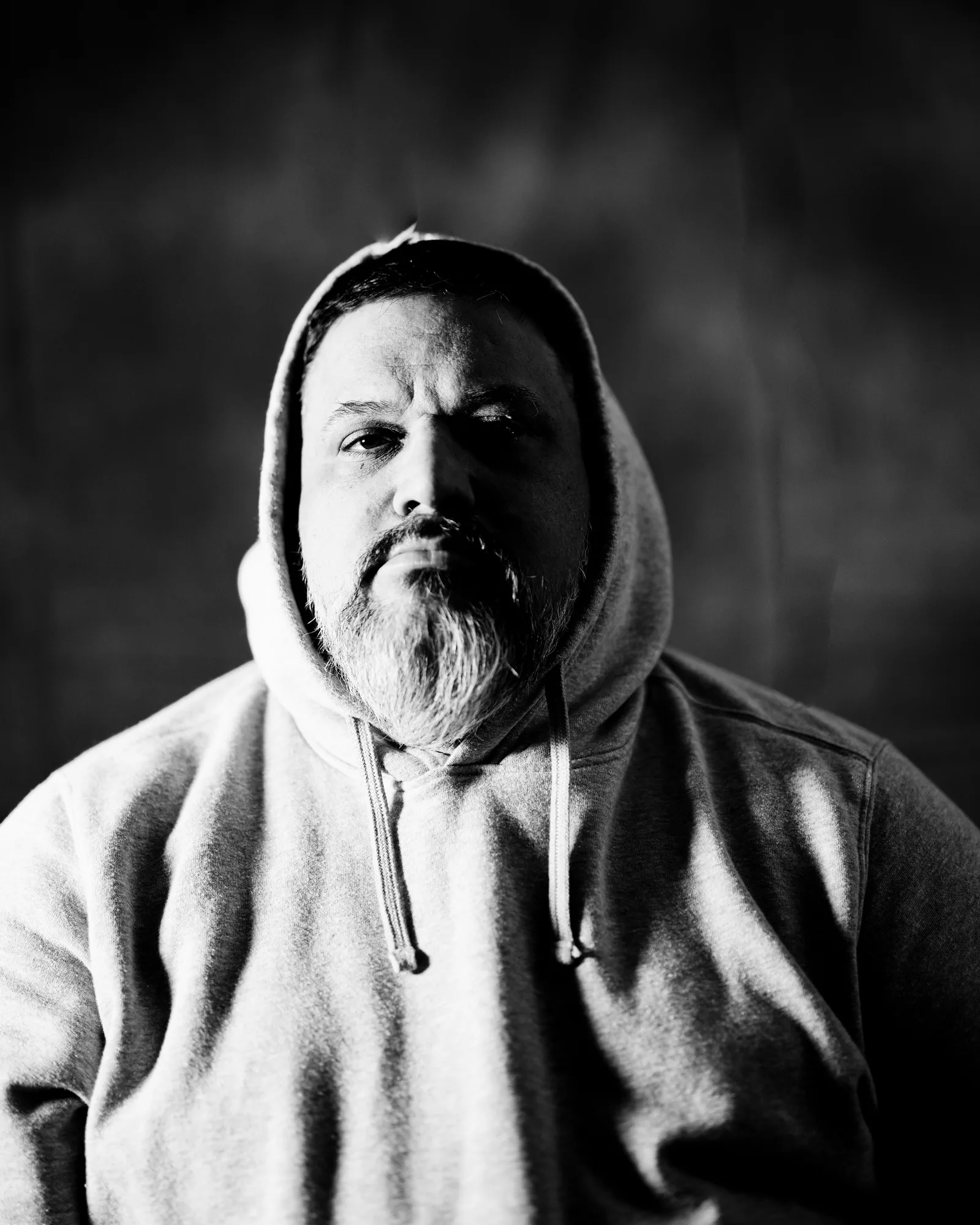Black-and-white close-up of a bearded man in a gray hoodie, head tilted down so only his beard and chest fill the frame.
