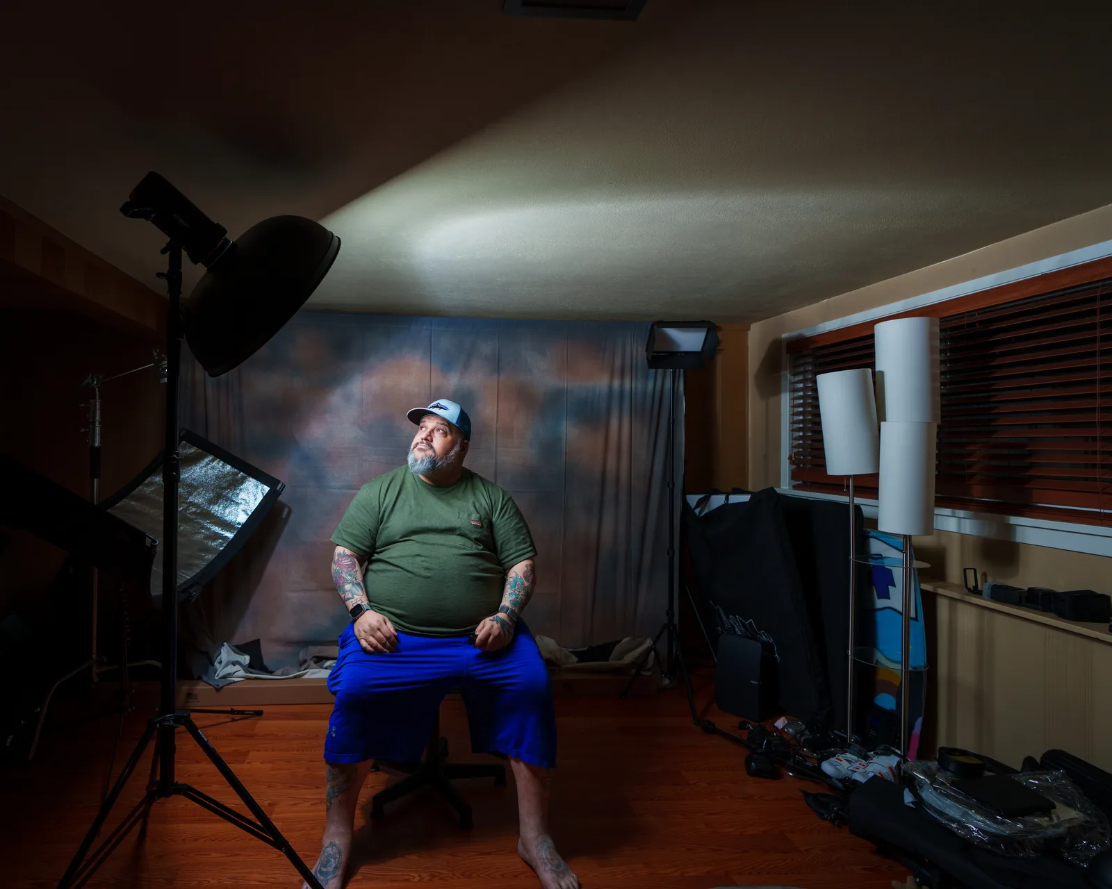 Bearded man in green t-shirt and white cap seated on a studio set, looking upward, surrounded by lights, backdrop, and window blinds.