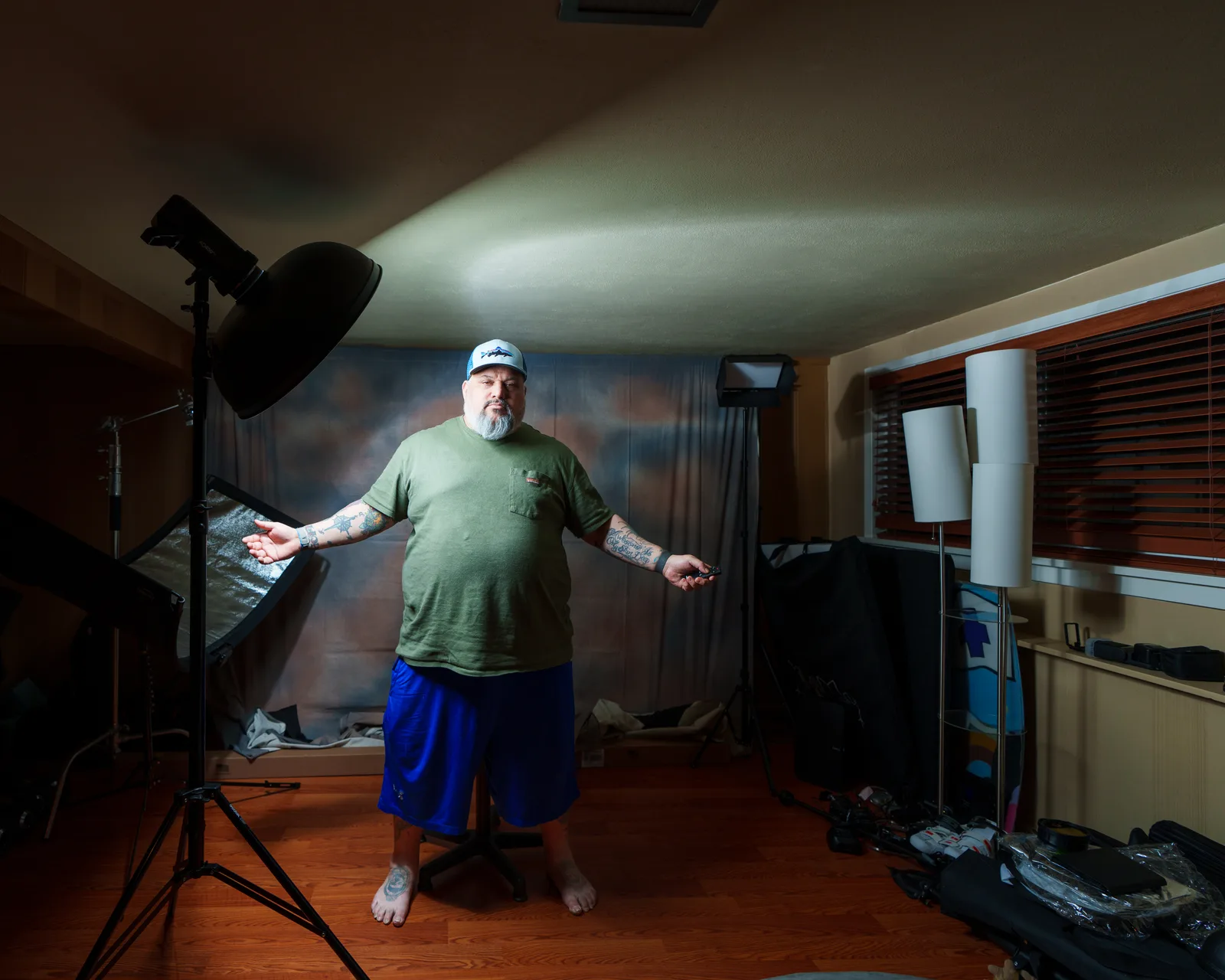 Bearded man in green t-shirt, blue shorts and ball cap stands arms open in home photo studio with lights and backdrop.