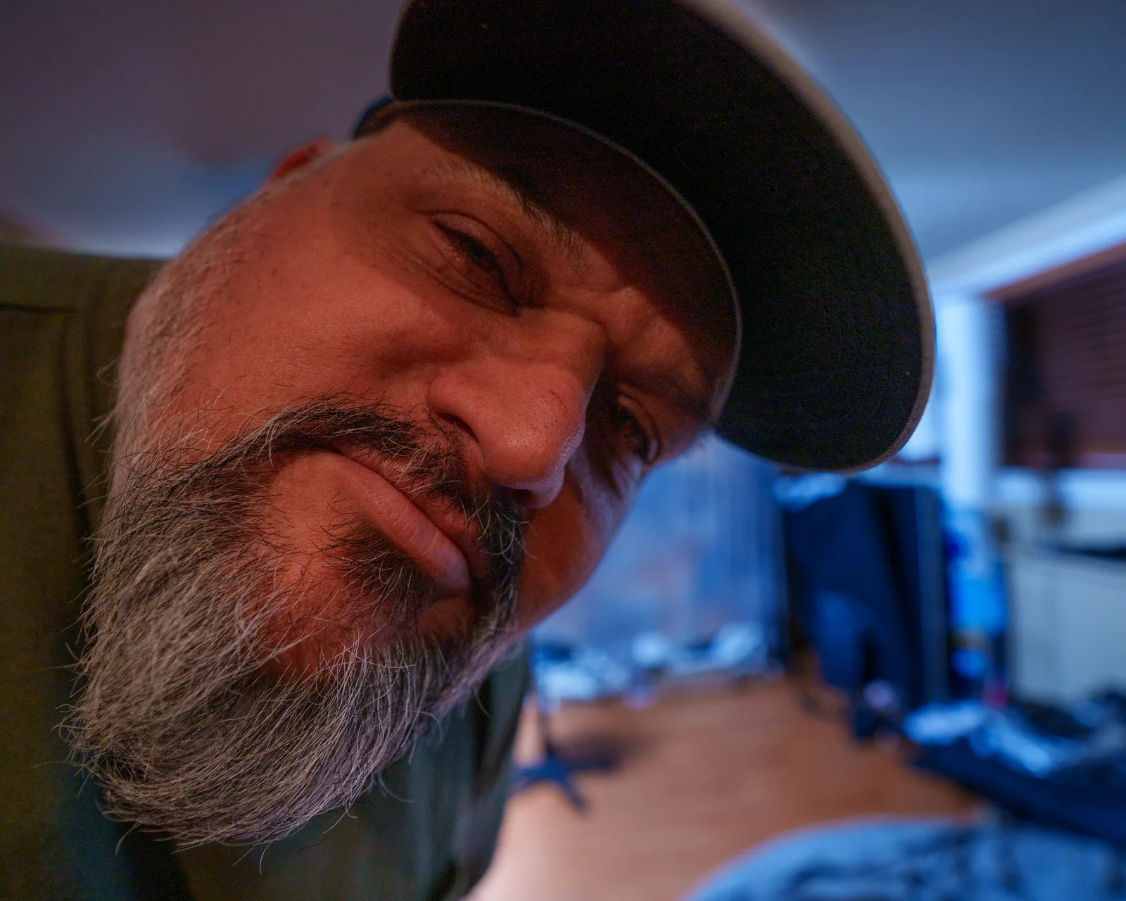 Extreme close-up of a bearded man in a dark cap tilting his face toward the camera, warm skin tones against a blue-lit room.