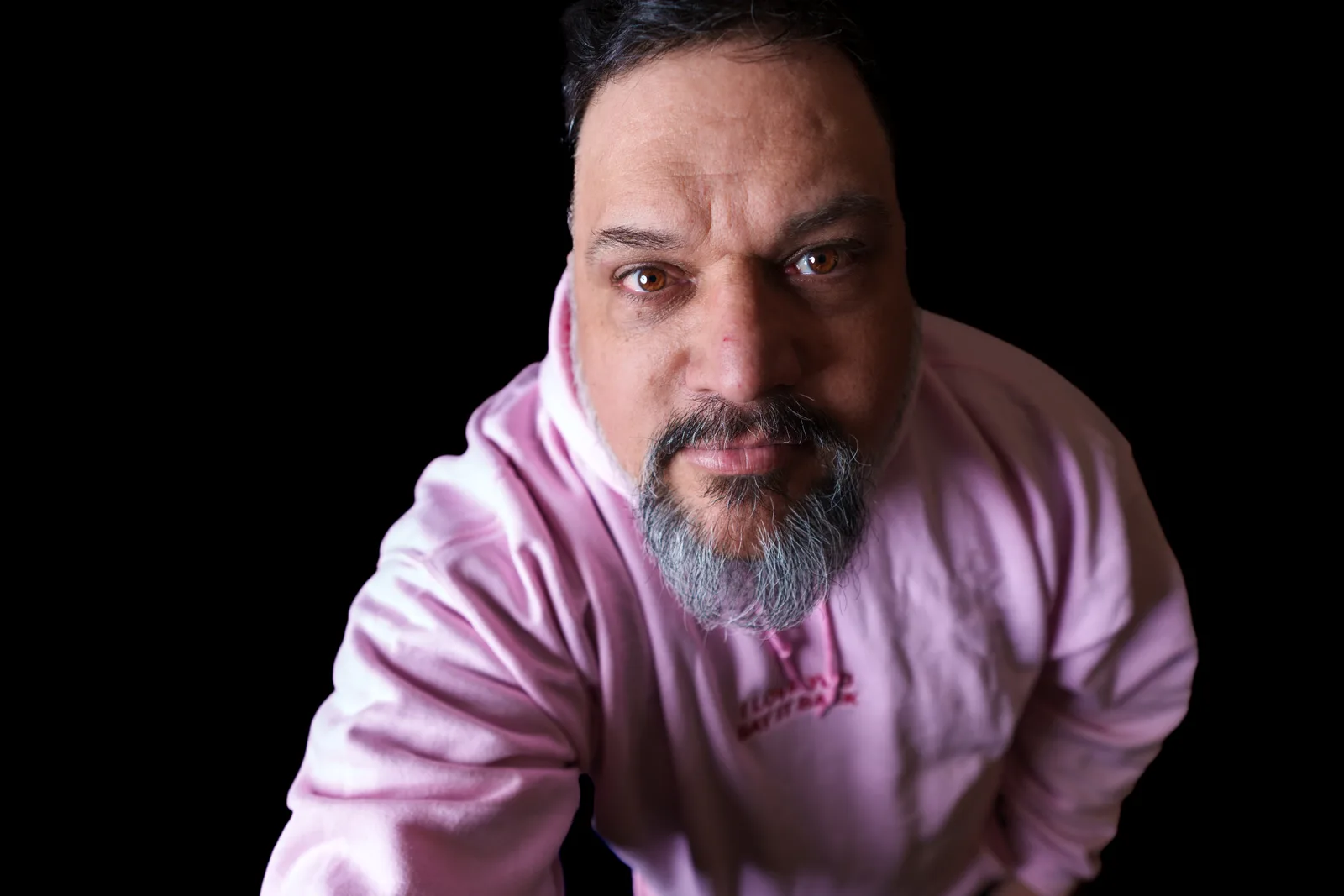 Bearded man in a pink hoodie photographed from above against black, looking up into the camera with calm eyes.