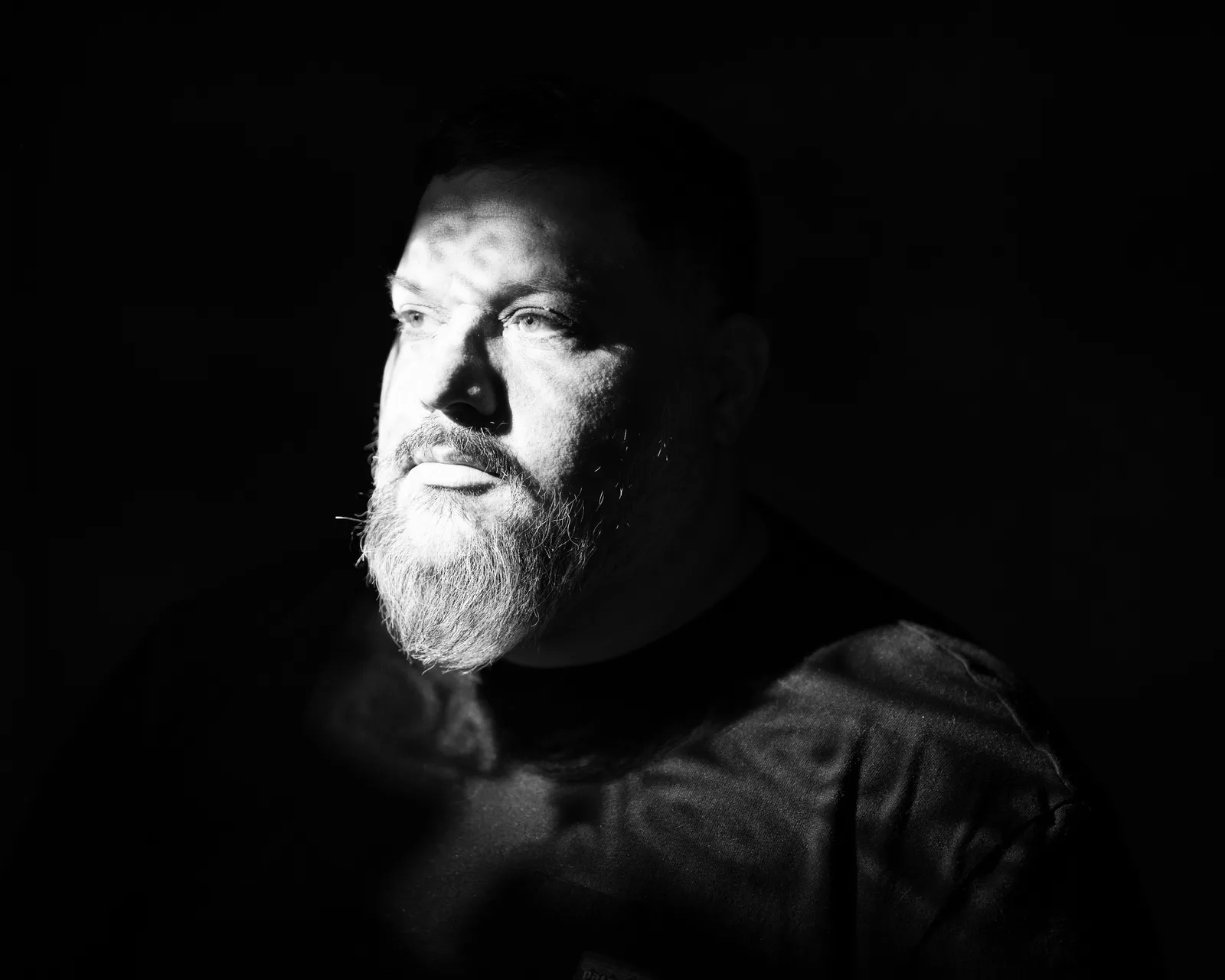 Black-and-white side-lit portrait of a bearded man against a dark background, gazing off-camera with a serious expression.