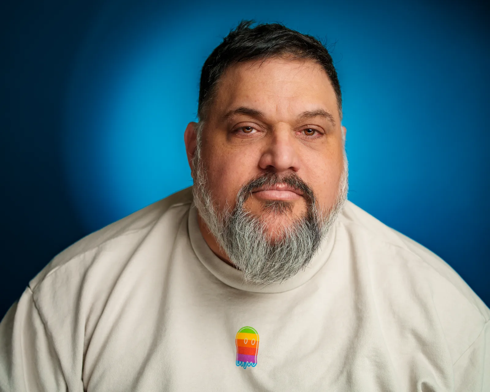 Close-up portrait of a bearded man in a cream t-shirt against a deep blue backdrop, gazing steadily at the camera.