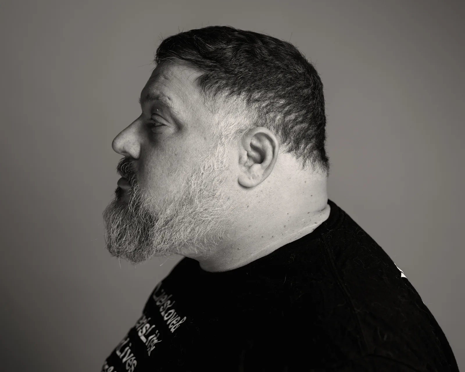 Black-and-white side profile of a bearded man with short hair, mouth slightly open, wearing a dark shirt against a plain backdrop.