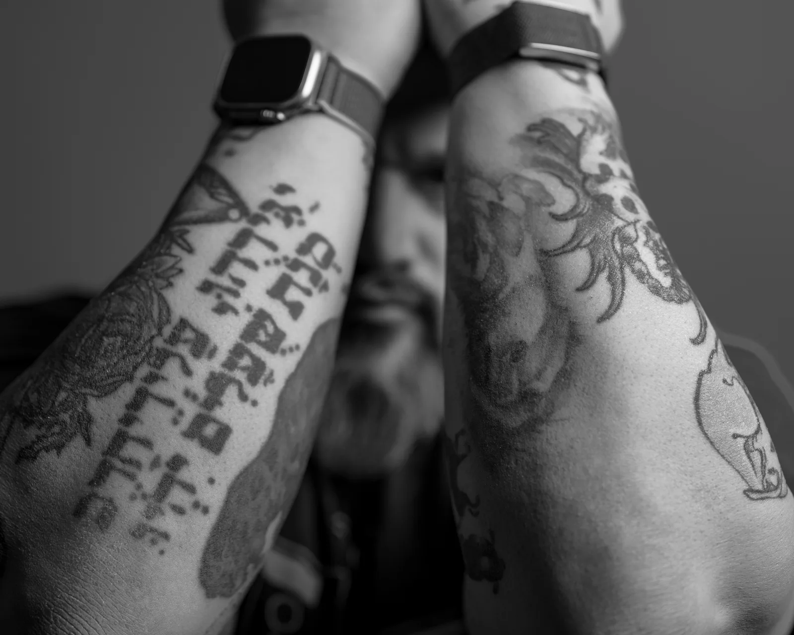 Black-and-white close-up of two heavily tattooed forearms raised in front of a bearded man's blurred face, smartwatch on wrist.
