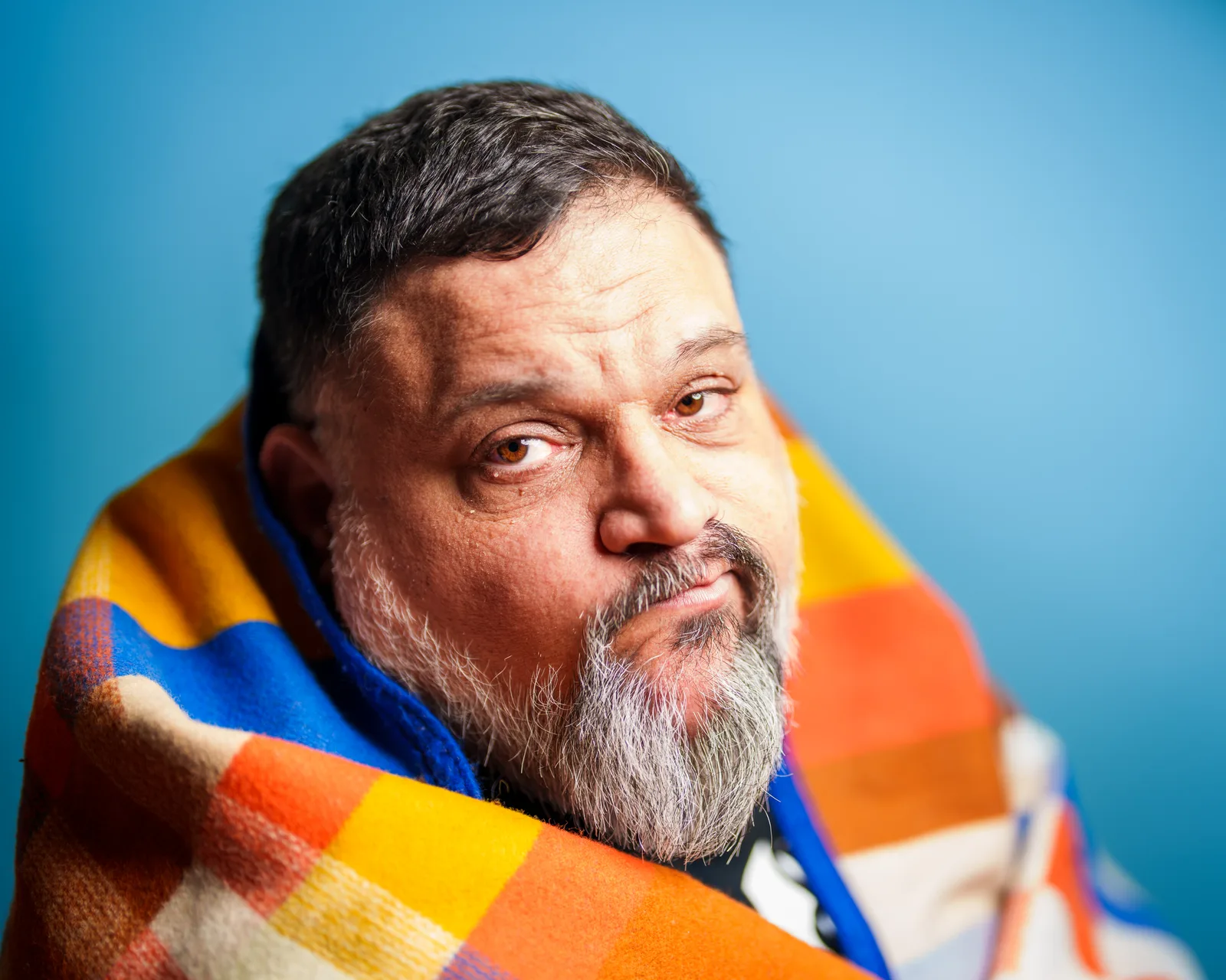 Bearded man with graying beard wrapped in a colorful orange, blue, and yellow plaid blanket, gazing sideways against a blue backdrop.