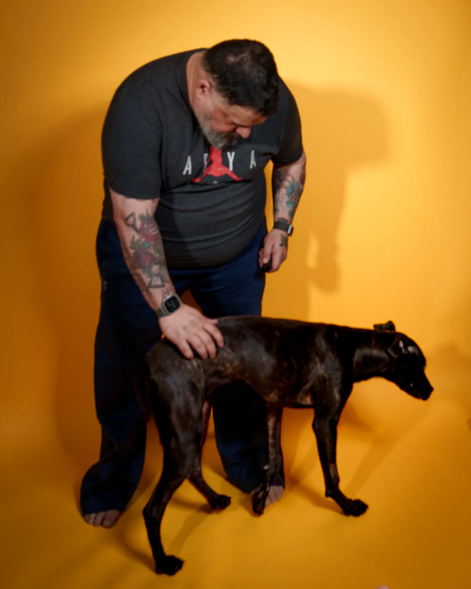 Bearded man in dark gray Air Jordan t-shirt bowing his head down, arms tattooed, against a bright yellow backdrop.