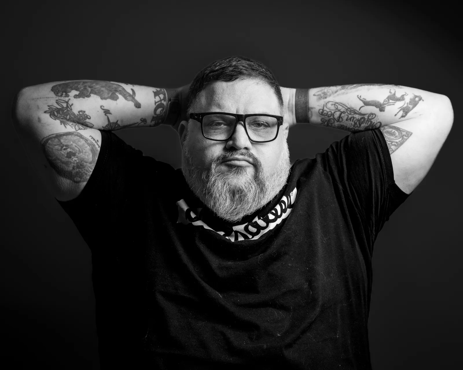 Black-and-white portrait of a bearded man in glasses and a black t-shirt, hands clasped behind his head, tattooed arms framing his face.