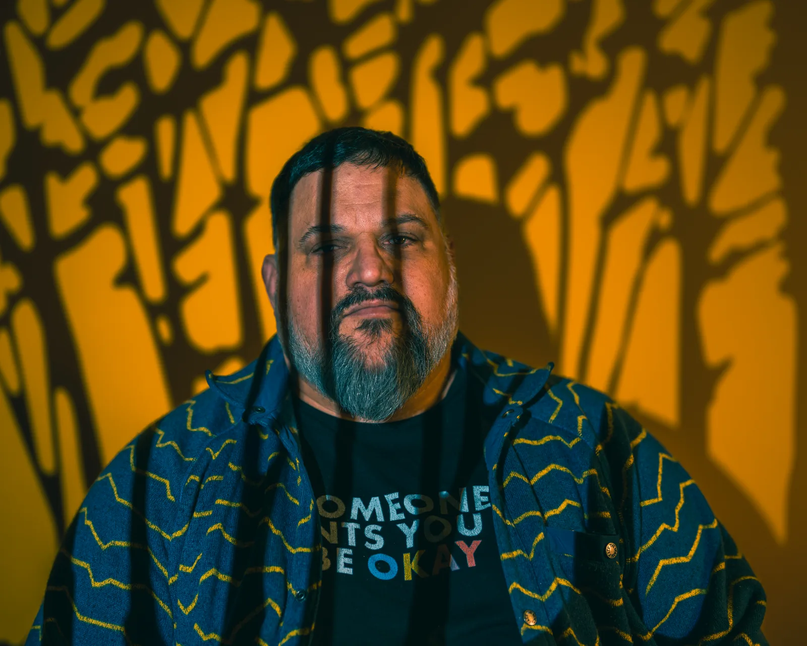 Bearded man's face lit in warm orange with dramatic branch-like shadows falling across him and the wall behind