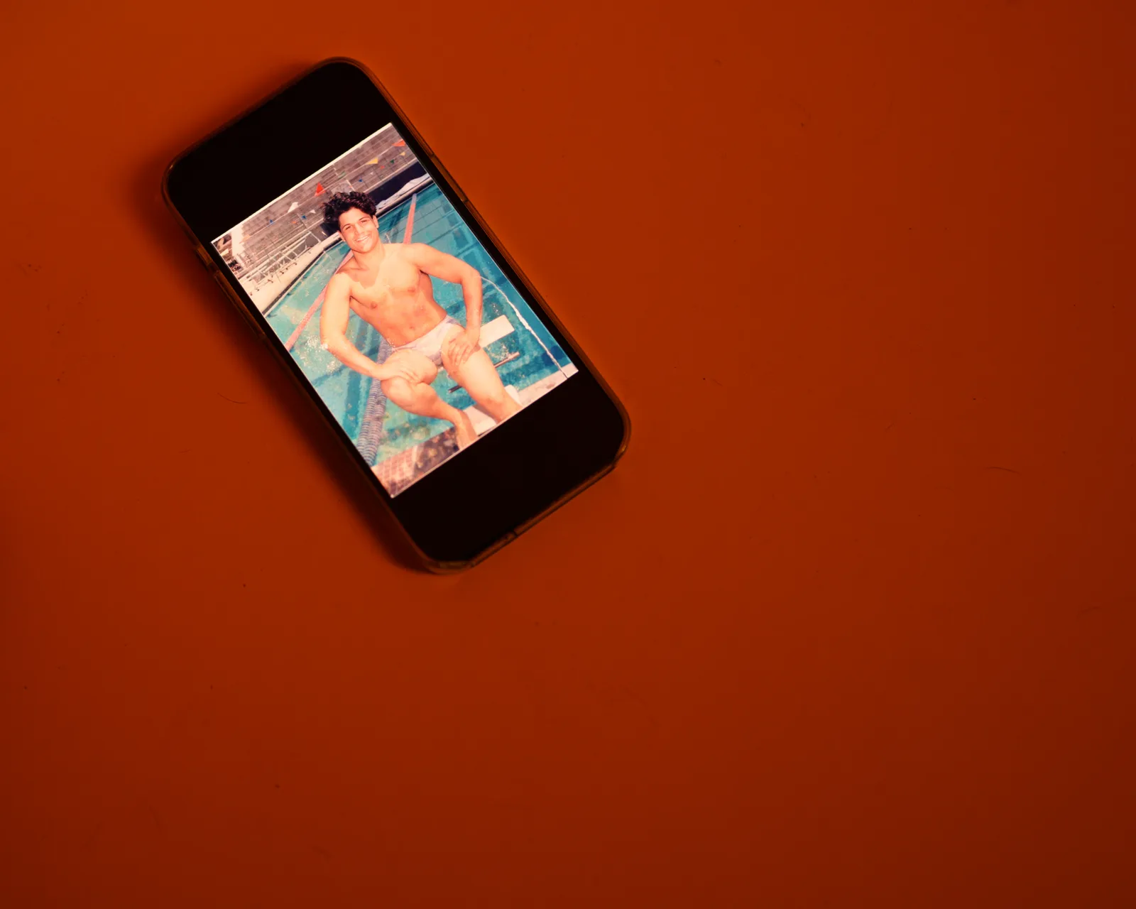 Smartphone on orange background showing an old photo of a young shirtless man in swim briefs posing by a pool.
