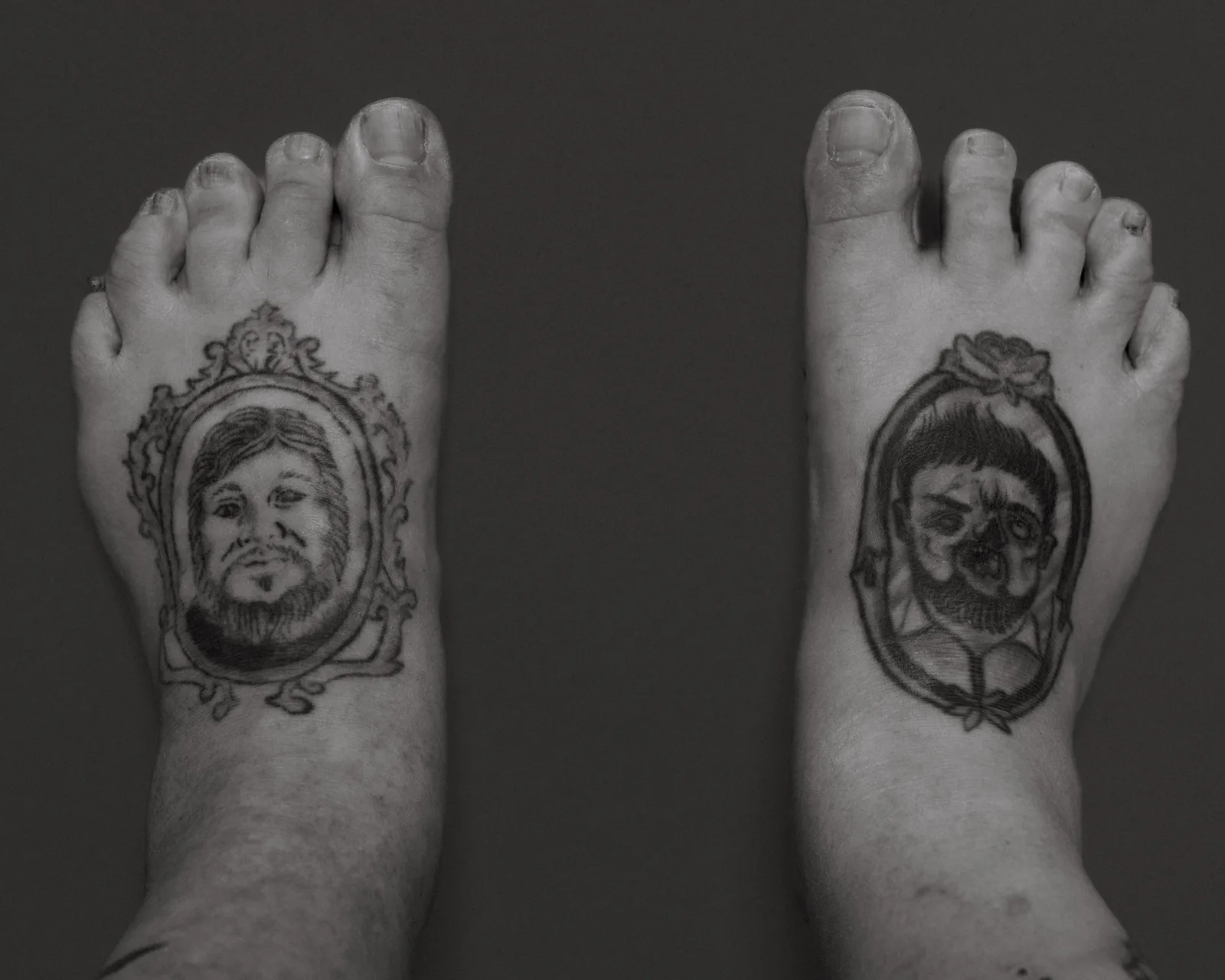 Black-and-white overhead view of two bare feet on dark background, each with a framed portrait tattoo: a bearded man and a zombie face.