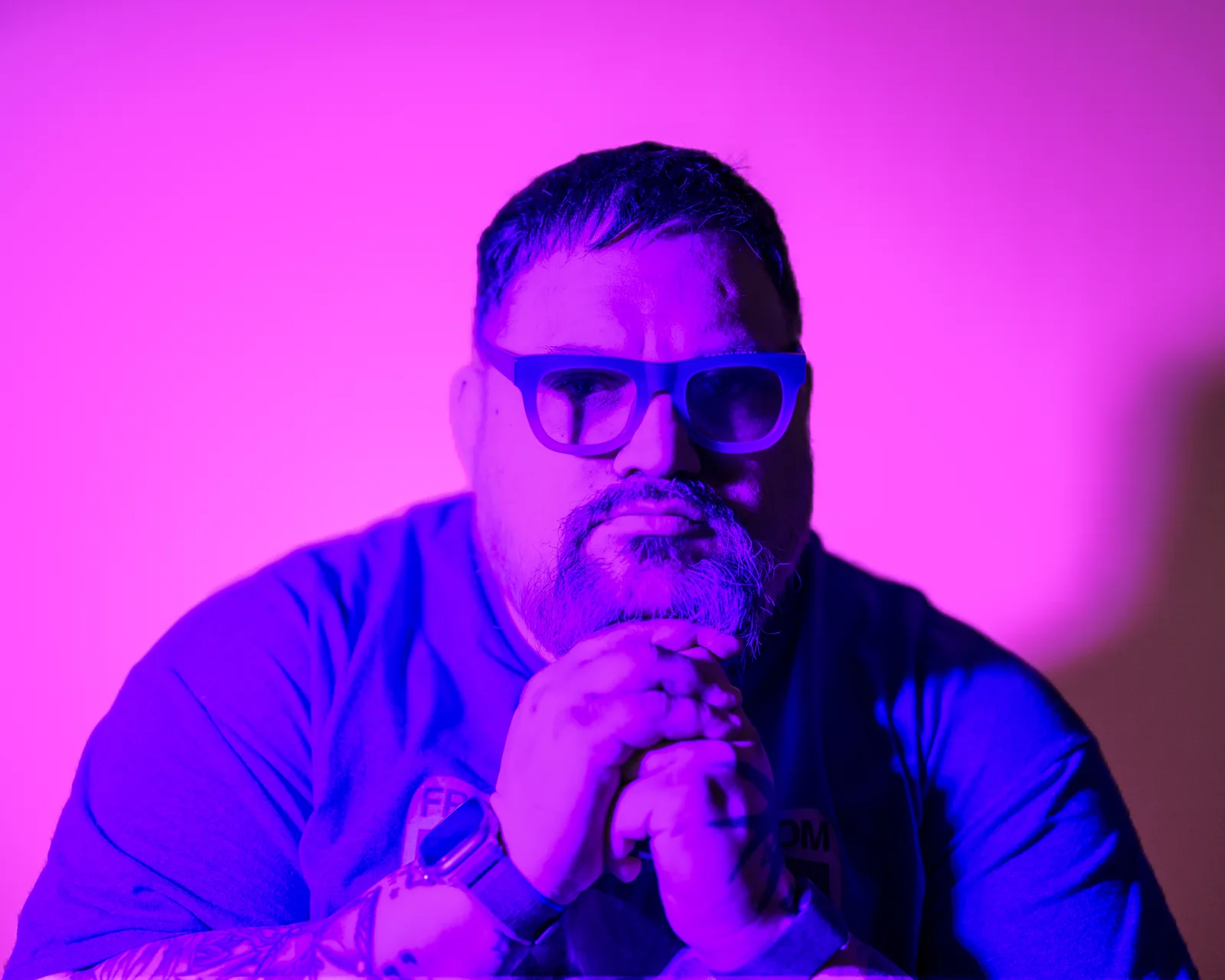Bearded man in blue glasses and shirt, hands clasped near chin, lit by saturated pink and purple light against a magenta backdrop.