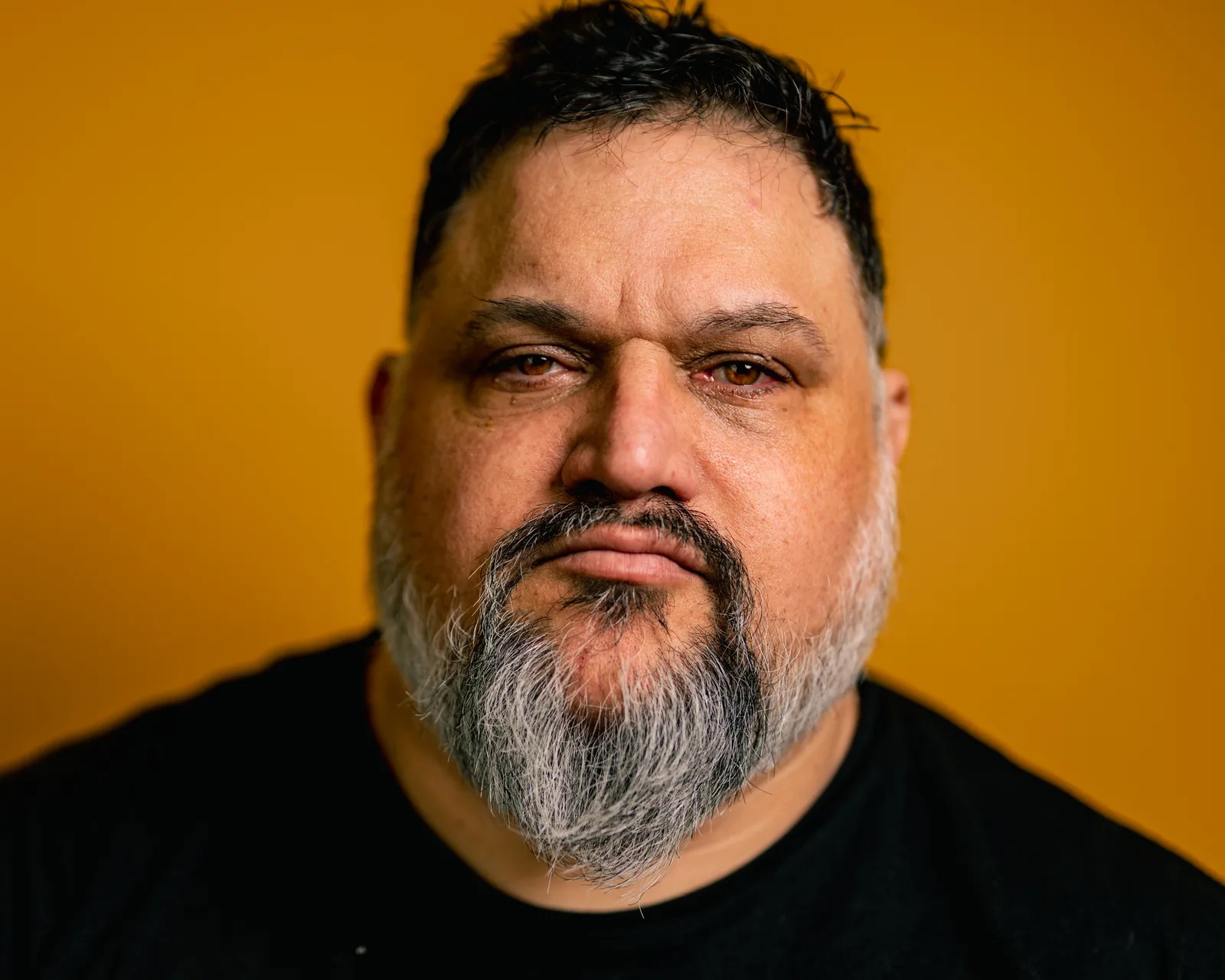 Tight close-up of a bearded man's face against a warm orange background, dark eyes looking directly at camera, neutral expression.