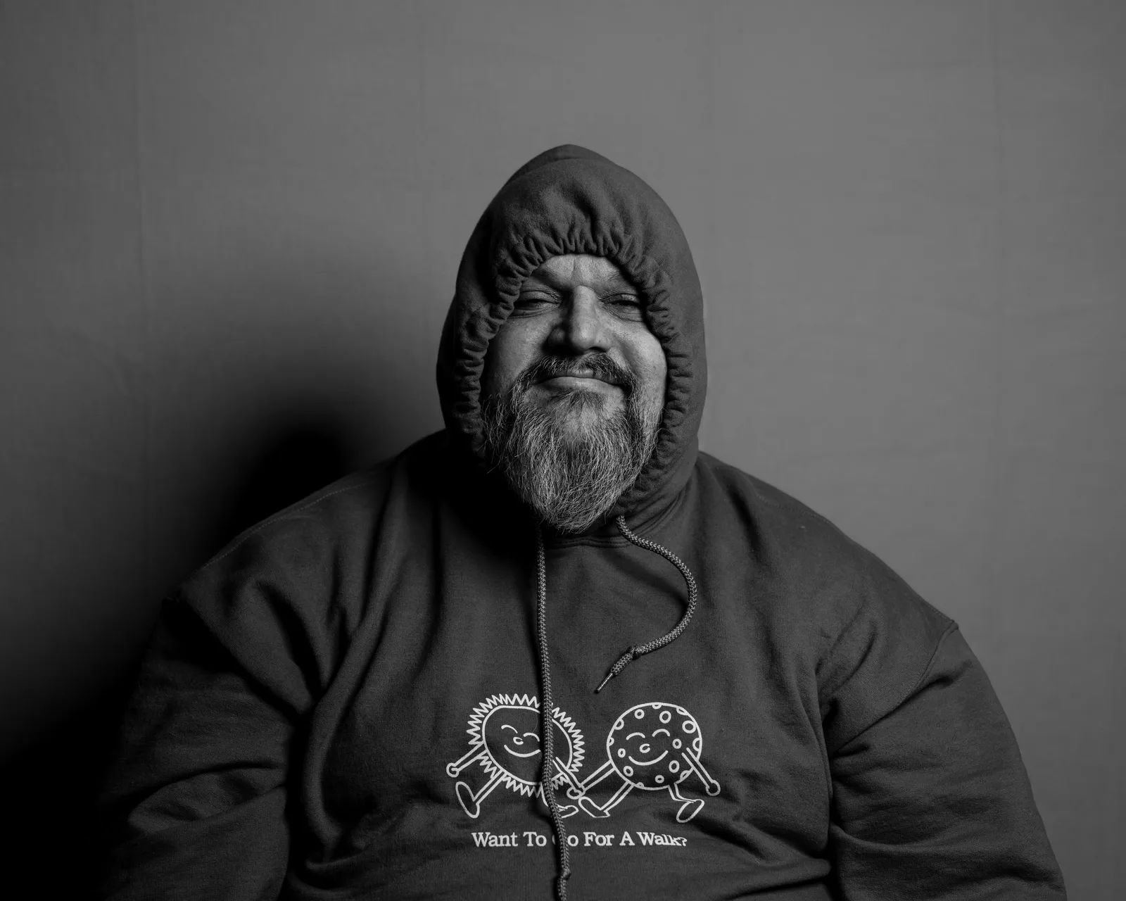 Black-and-white portrait of a bearded man in a dark hoodie pulled up over his head, smirking slightly at the camera.