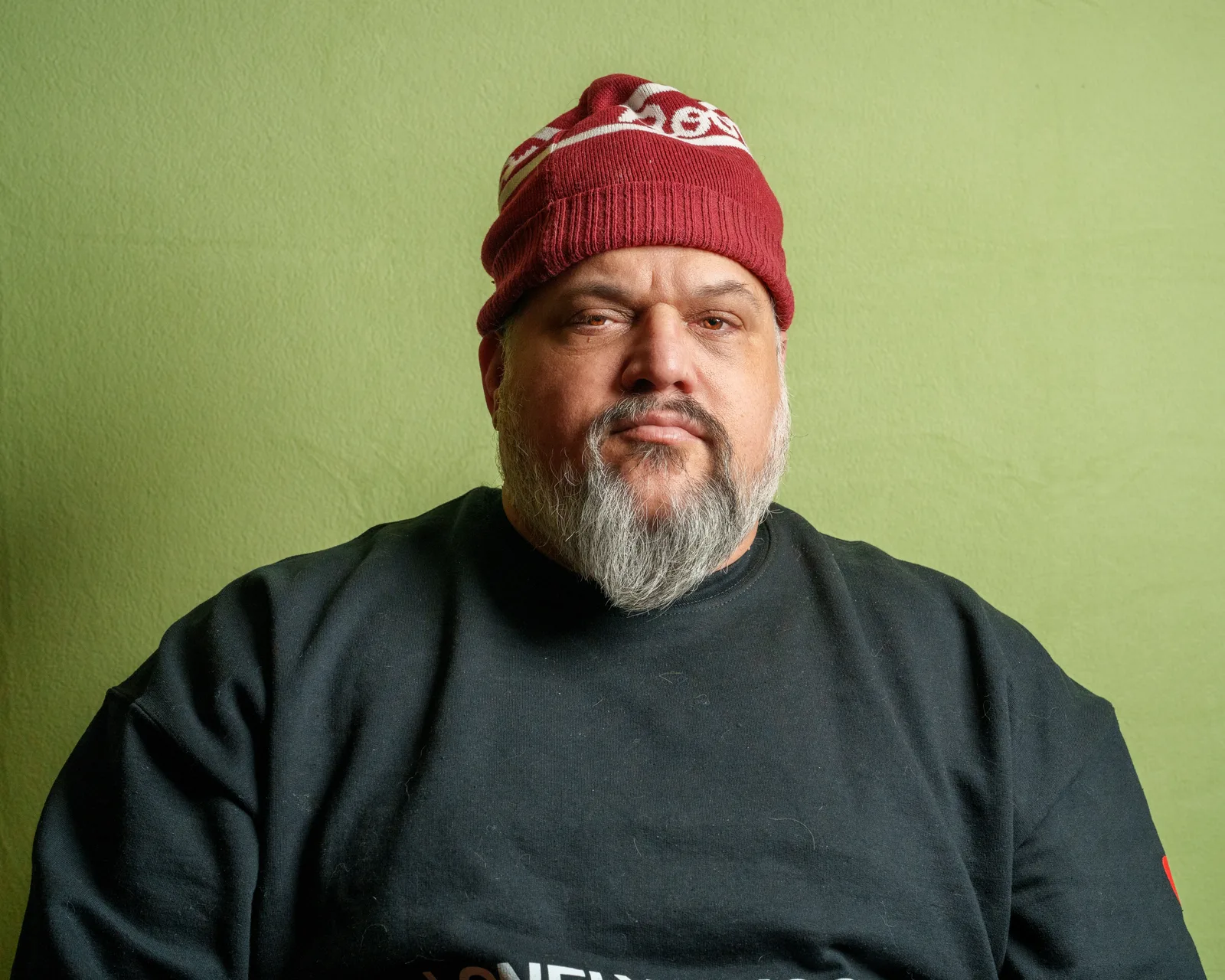 Bearded man in a red knit beanie and dark t-shirt against a green wall, heavy-lidded eyes, weary expression.