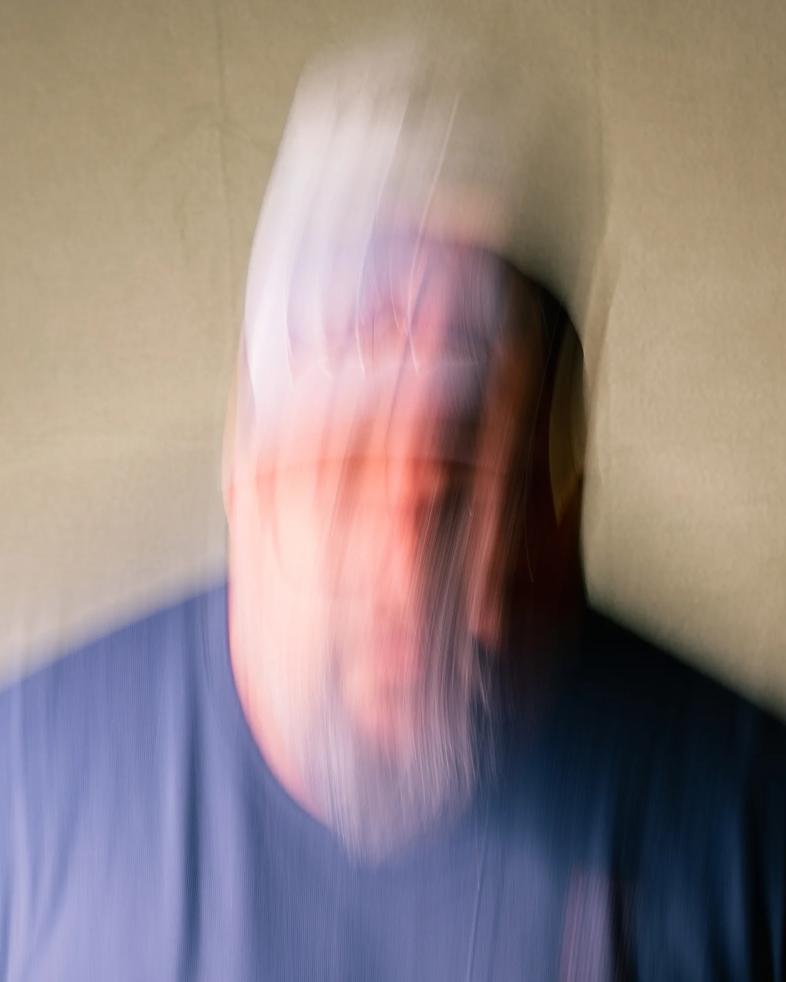 Heavily motion-blurred self-portrait, face streaked into smears of pink and orange against a blue shirt and pale wall.