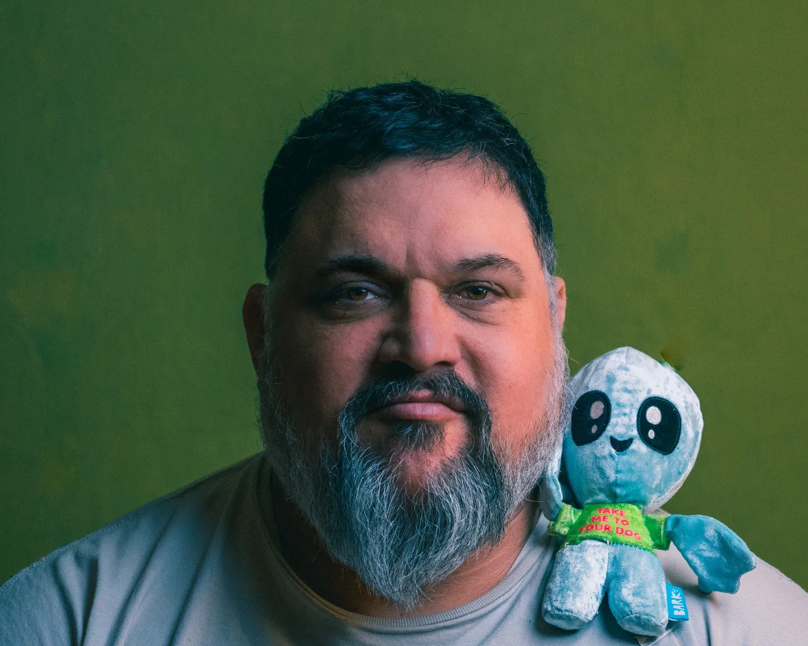 Close-up portrait of a bearded man with graying goatee against a green wall, holding a small blue plush alien that reads "Take me to your dog" near his shoulder.