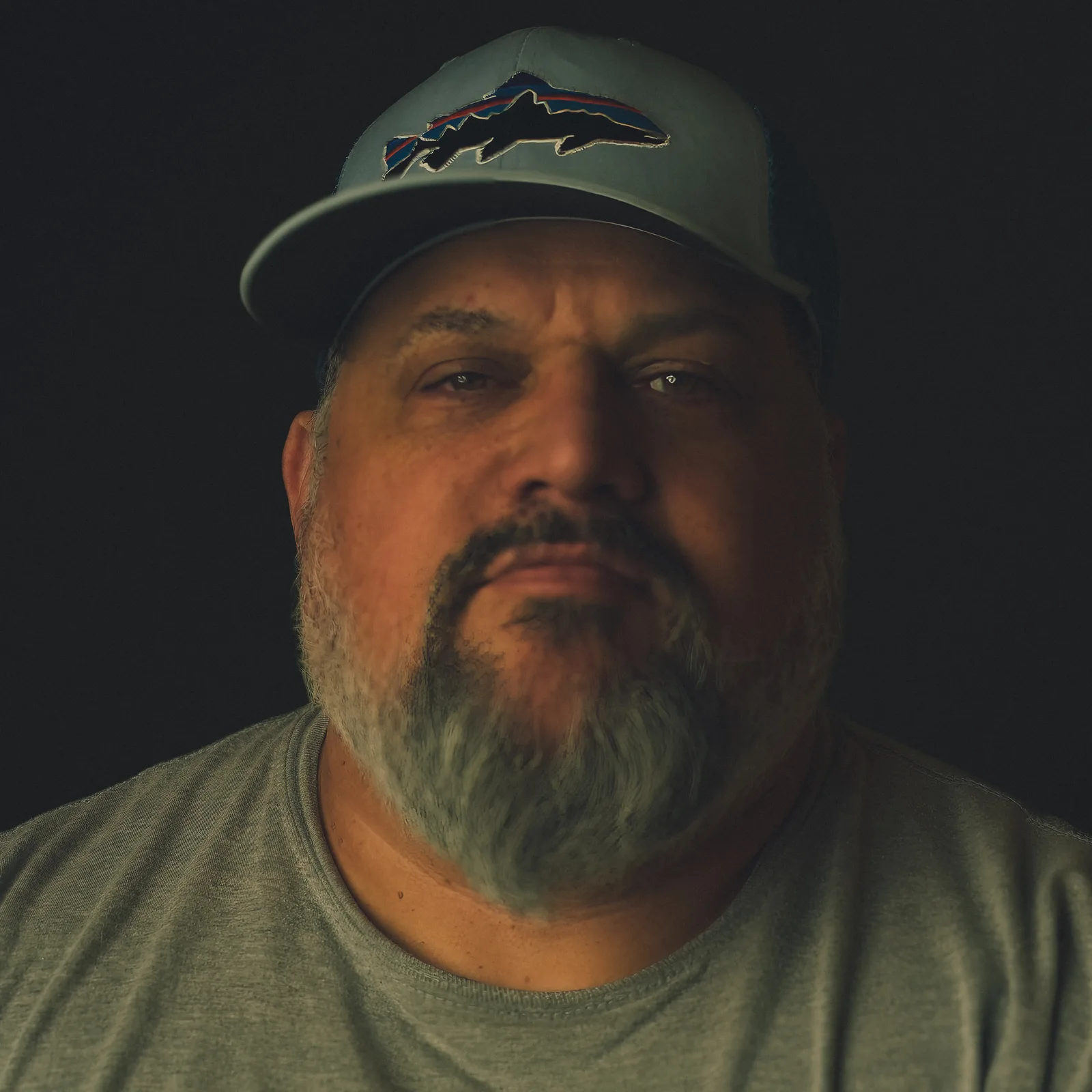 Tight crop on a man's gray bearded chin and mouth above a heather t-shirt, face dimly lit against black.