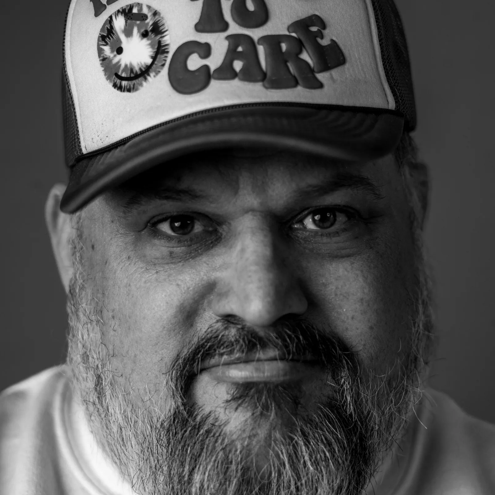 Black-and-white tight crop on a man's eyes and white trucker hat reading 'TO CARE' with a tie-dye smiley patch.