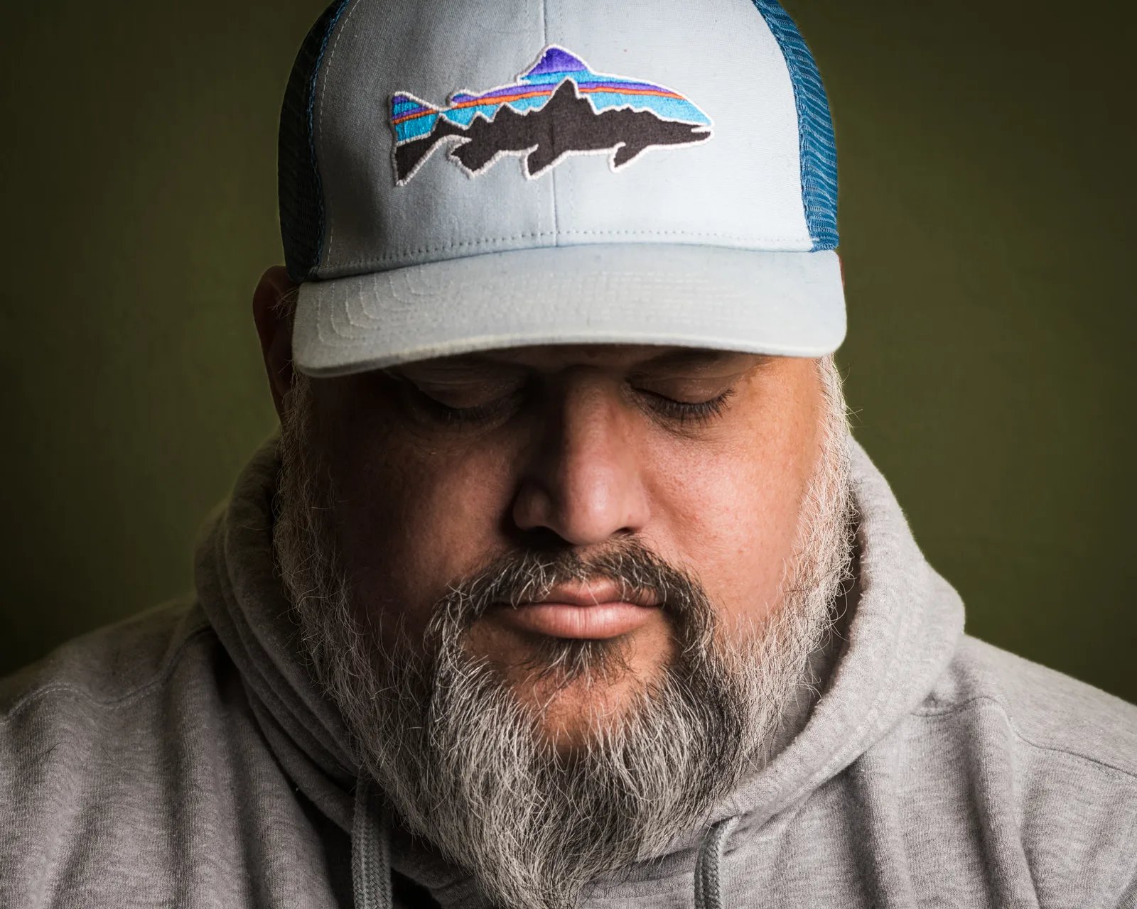 Close-up of a bearded man in a gray hoodie and pale cap, eyes downcast, against a dark olive background.