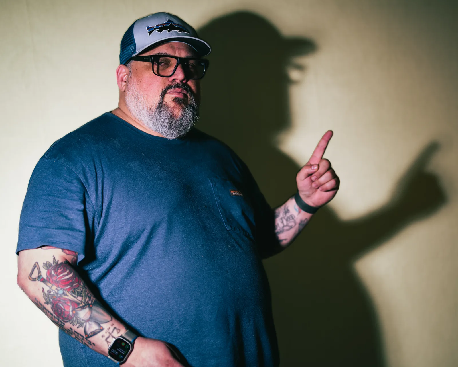 Bearded man in navy t-shirt pointing upward with one finger, tattoos on forearm, smartwatch on wrist, his shadow cast on wall behind