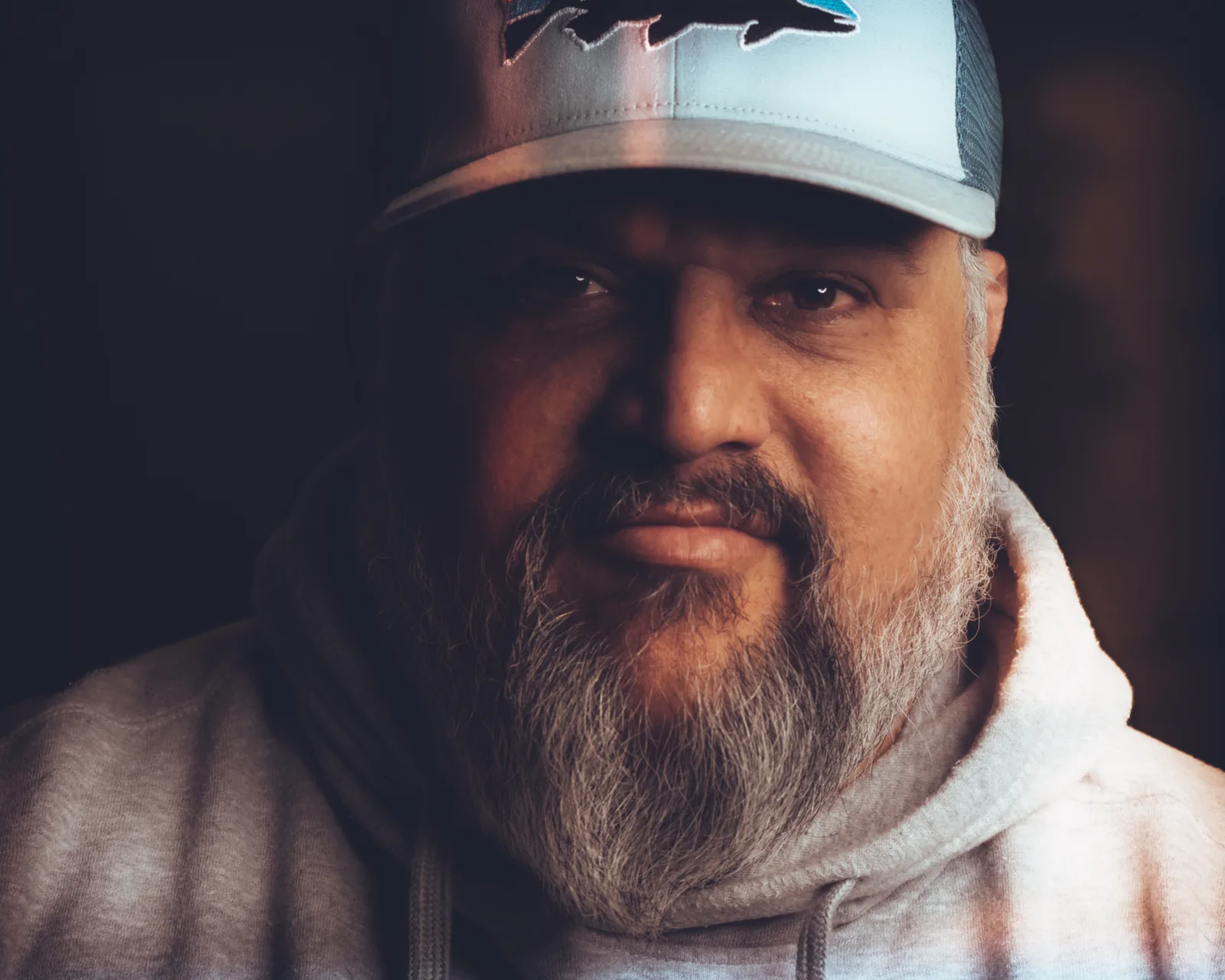 Tight close-up of a bearded man's lower face and mouth, wearing a light gray hoodie, lit warmly against a dark background.