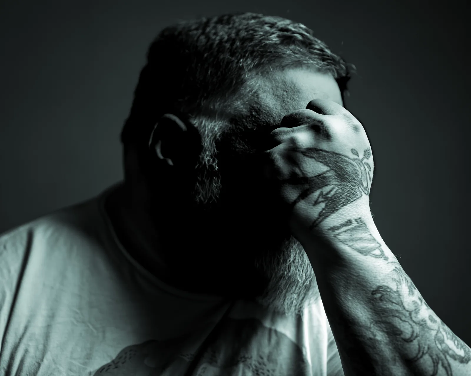 Black-and-white close crop of a bearded man in a white t-shirt, head bowed into a heavily tattooed hand covering his face.
