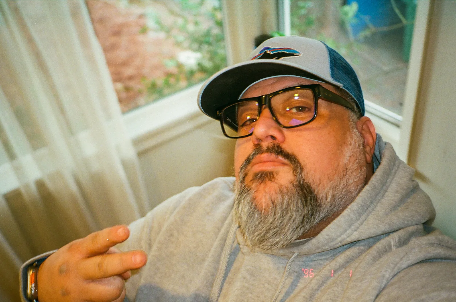 Bearded man in glasses and gray trucker cap looks up at camera, wearing gray hoodie, near sheer-curtained window