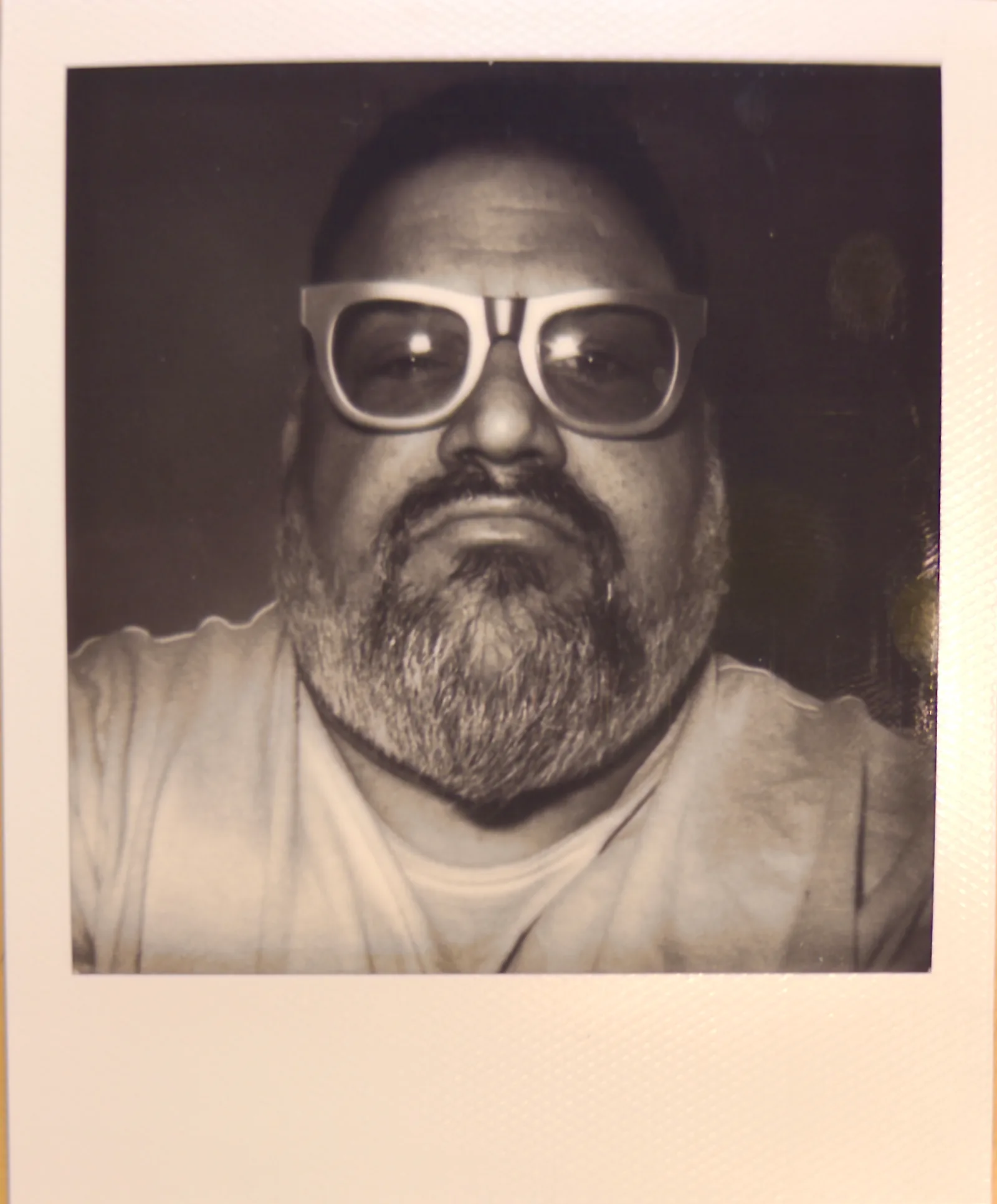 Sepia-toned instant photo, close-up of a bearded man in chunky white-framed glasses, only forehead, eyes and mustache in frame.