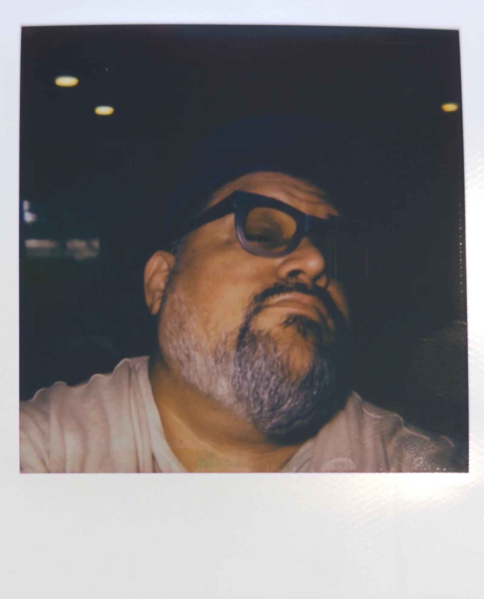 Tight low-angle close-up of a bearded man in dark glasses and a cap, face lit warmly against deep shadow.