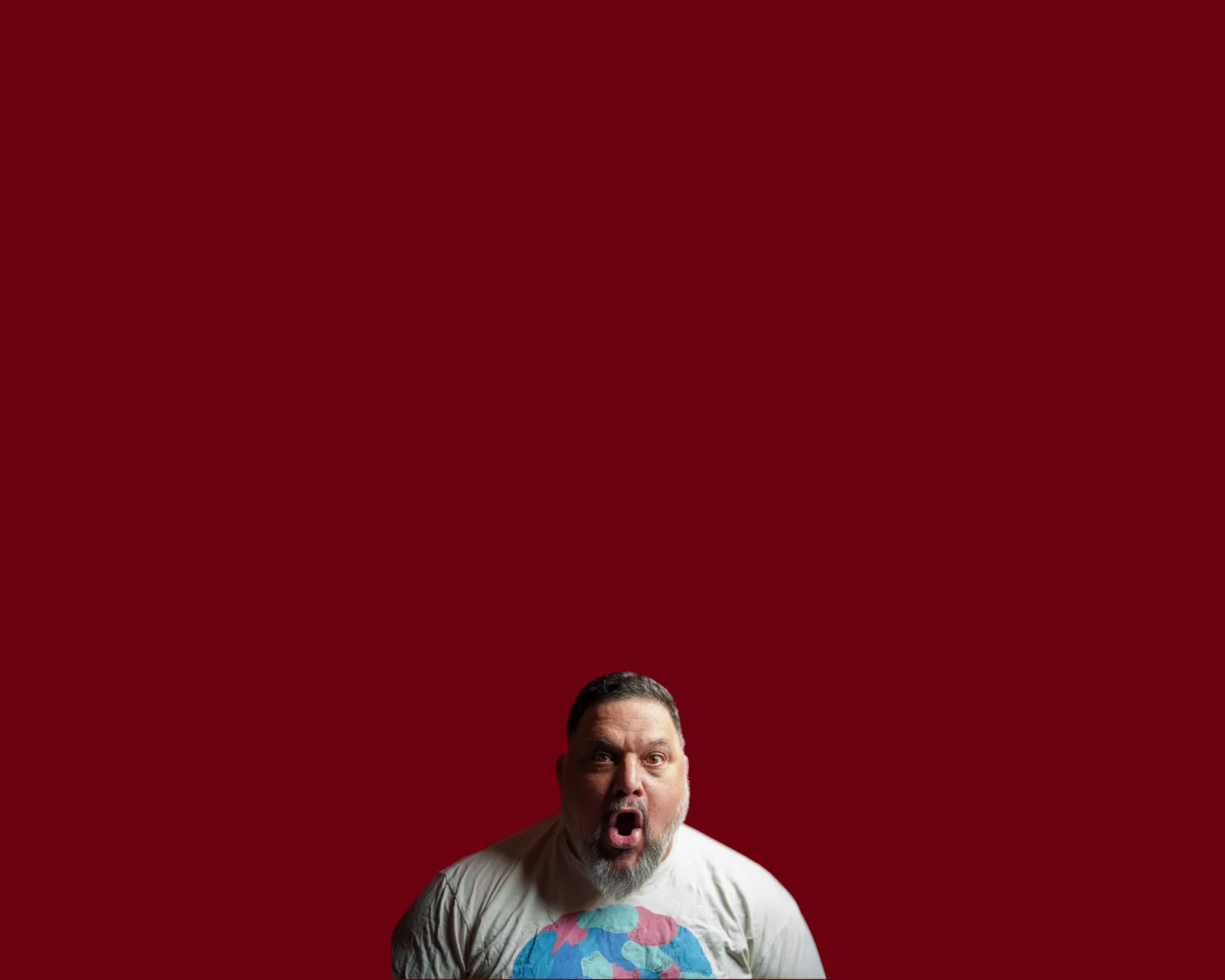 Man with graying beard and short hair, mouth wide open in shock or shout, wearing a white t-shirt with a globe graphic, against a deep red background.
