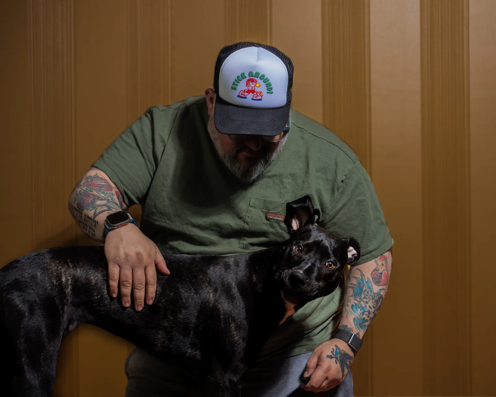 Bearded man in olive Patagonia tee and white trucker hat reading 'Stick Around', head bowed toward a black dog, tattooed arm visible