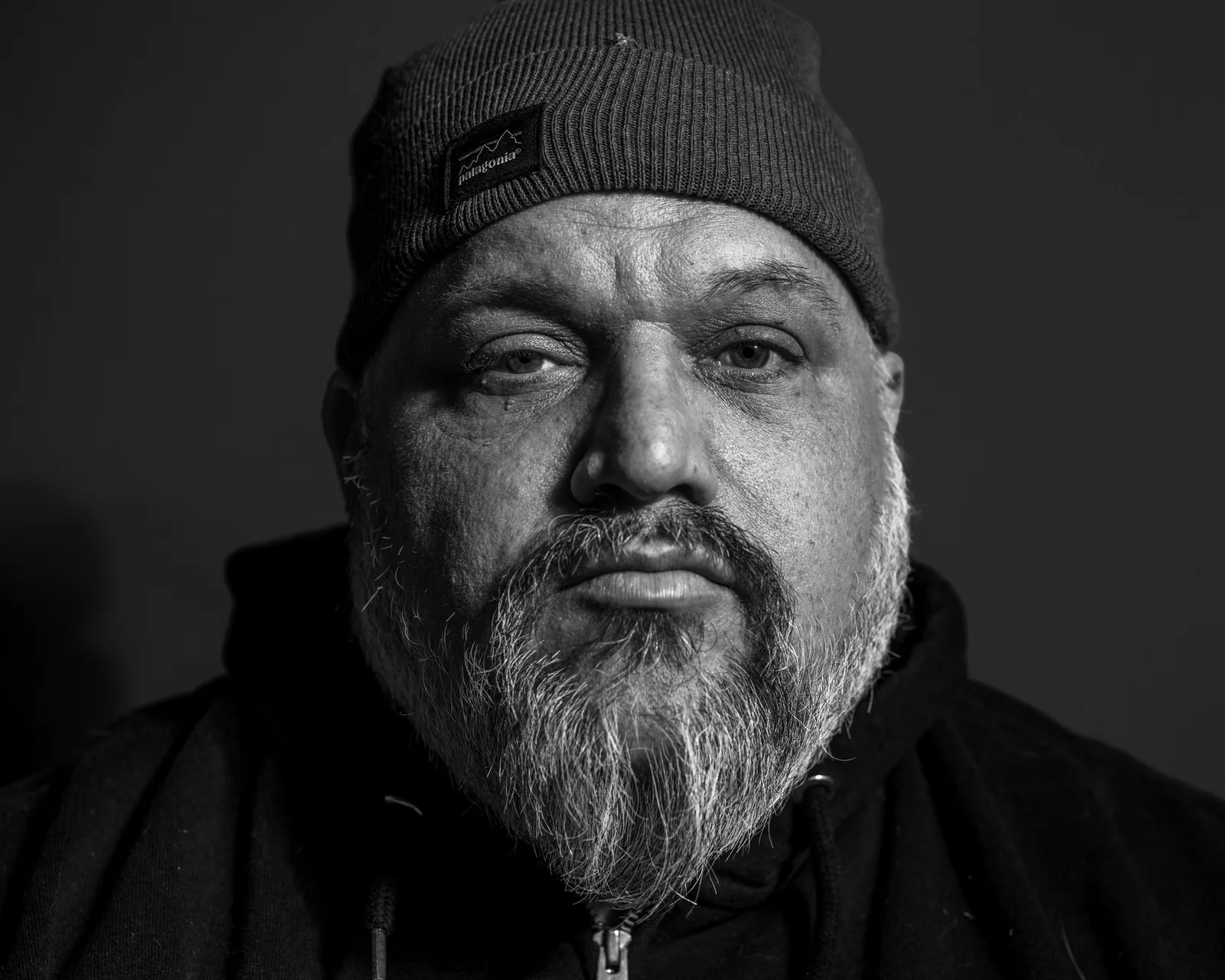 Black-and-white tight close-up of a bearded man in a Patagonia beanie, weary eyes looking directly at the camera.