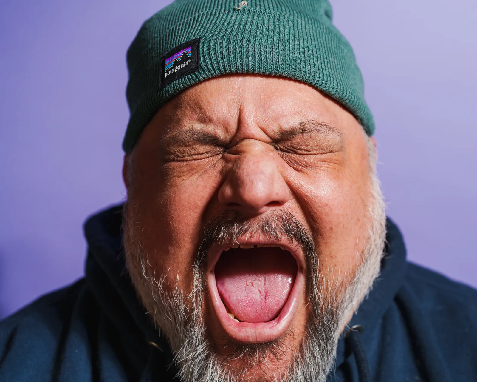 Tight close-up of a bearded man's face screwed up in a shout or grimace, eyes clenched shut, mouth wide open, wearing a green Patagonia beanie against a lavender backdrop.