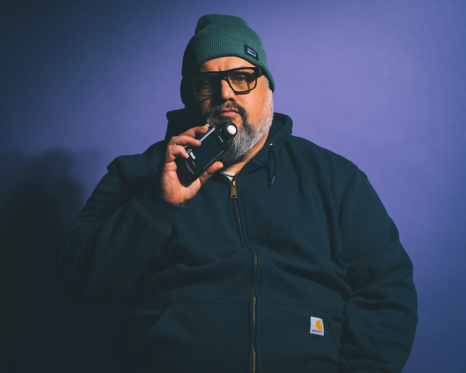 Bearded man in green beanie and dark hoodie holds a light meter to his chin, glasses on, against a purple backdrop.