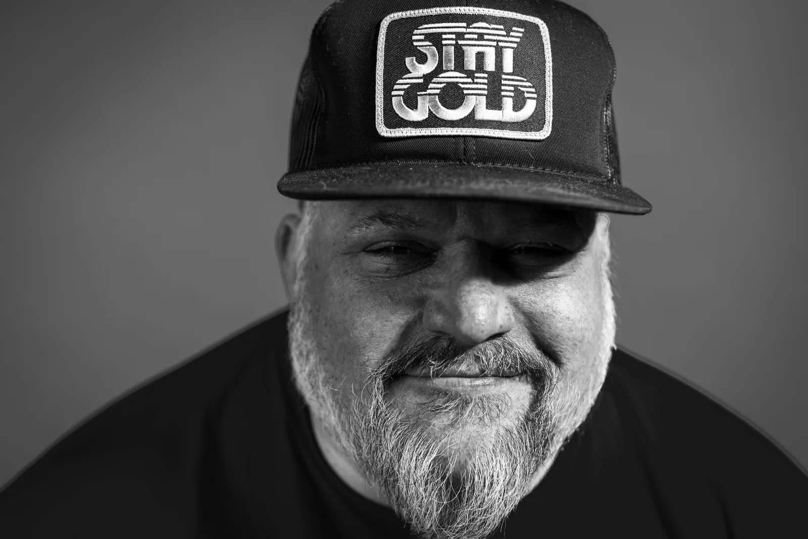 Black-and-white close-up portrait of a bearded man in a dark trucker hat and black shirt, smiling softly with downcast eyes.